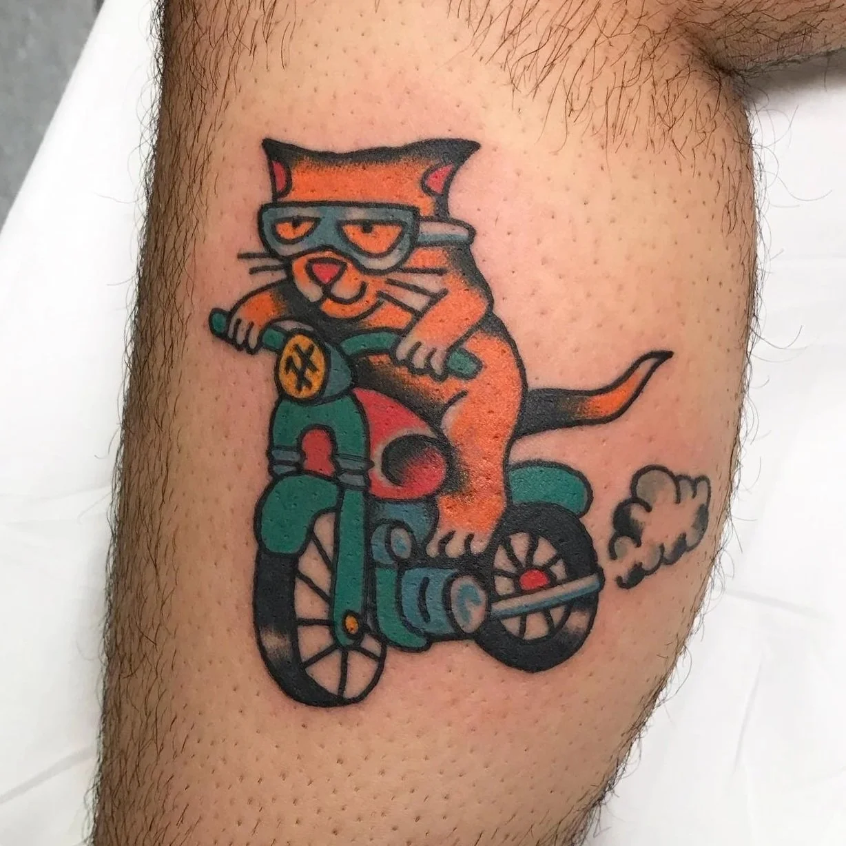 A colorful tattoo of an orange cat wearing goggles, riding a teal-colored motorcycle with a cloud of smoke behind.