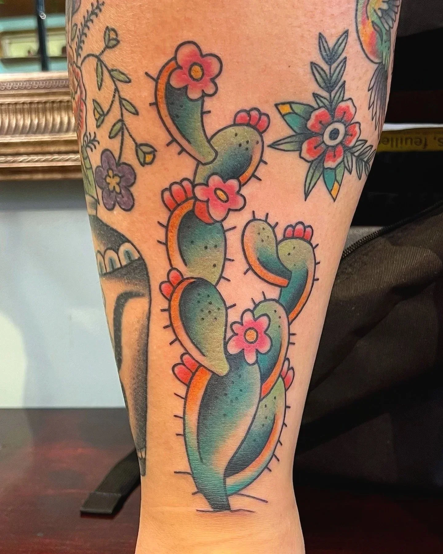 Colorful tattoo of a cactus with pink flowers and green pads, outlined in black, on a person's arm.