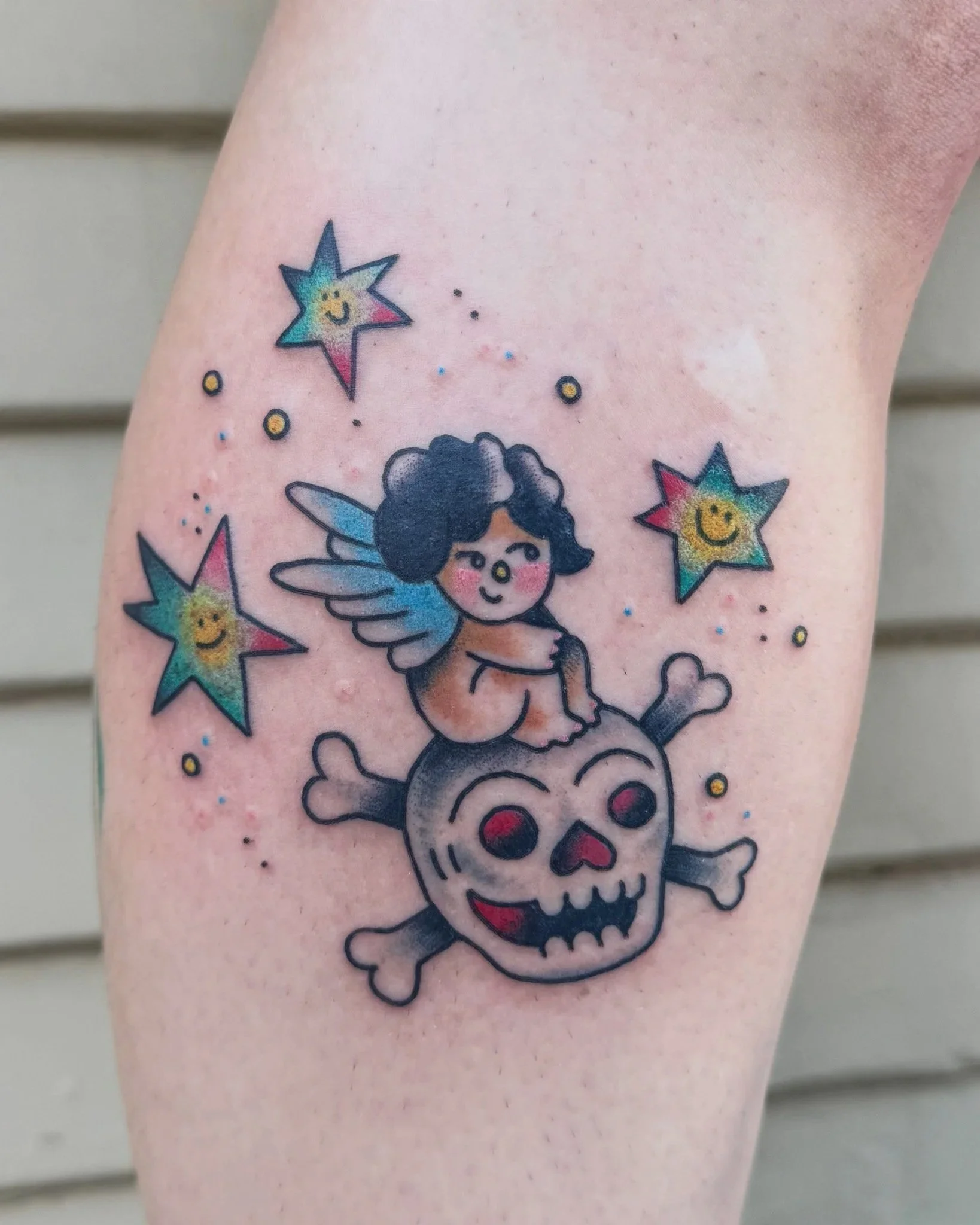 Colorful tattoo of a cherub sitting on a smiling skull with rainbow-colored stars and small dots around.