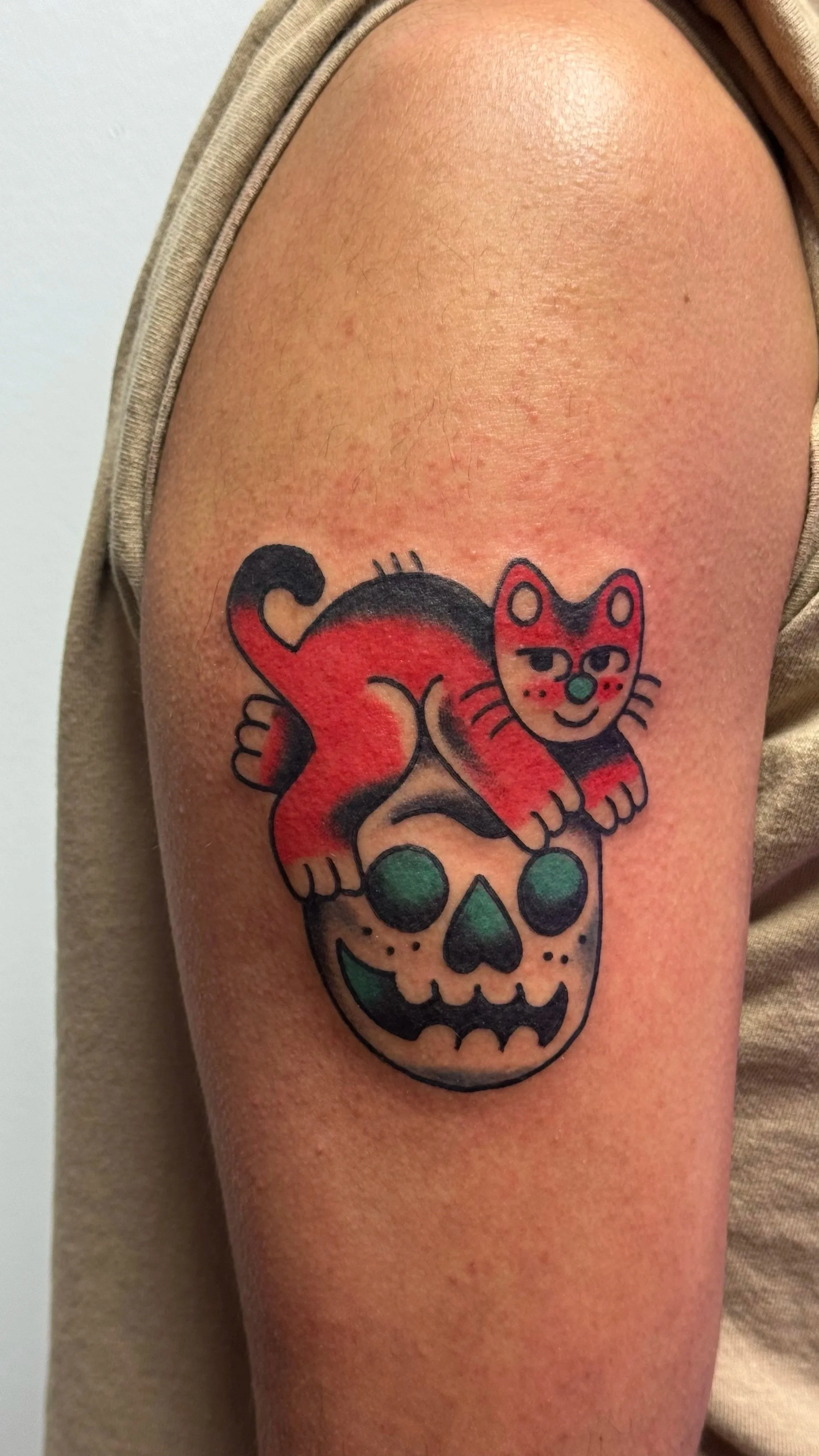 Tattoo of a red and black cat with a smiling face sitting on top of a green and black skull on a person's upper arm.