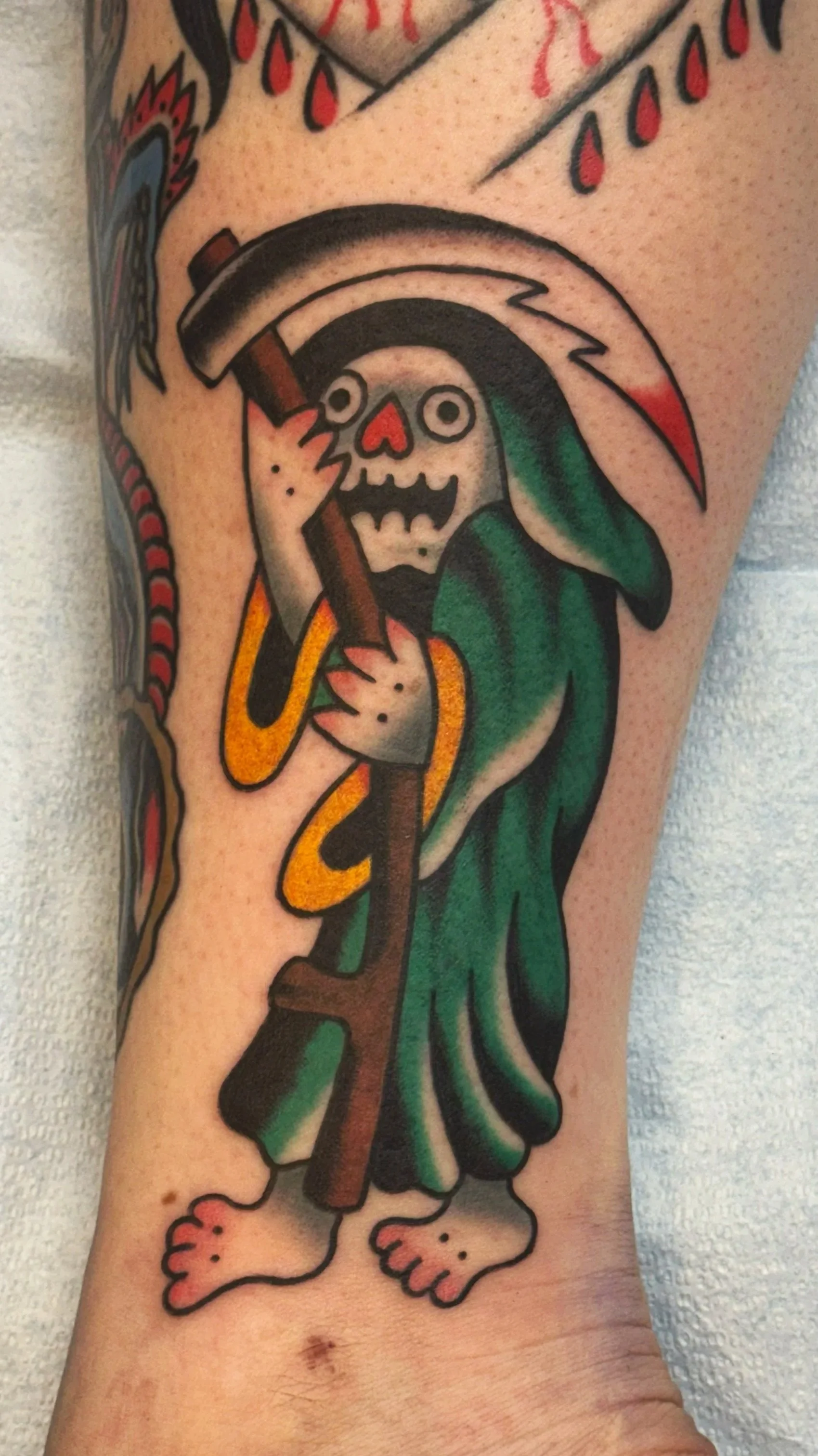 A tattoo of the Grim Reaper with a cartoonish skeleton face, wearing a green hooded cloak, holding a large scythe with a curved blade, and standing barefoot with tiny feet.