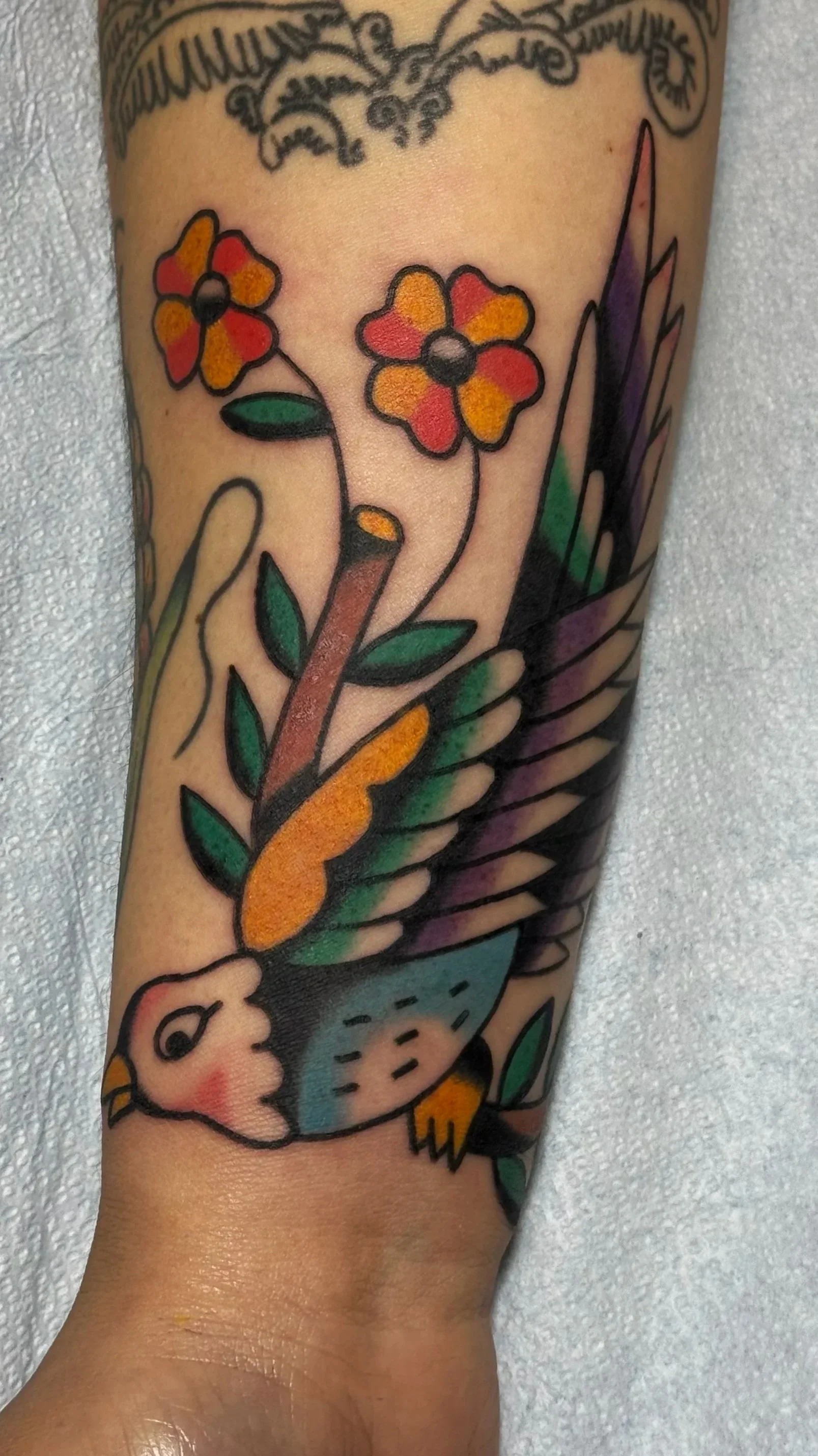 A colorful tattoo of a bird with outstretched wings holding flowers in its beak.