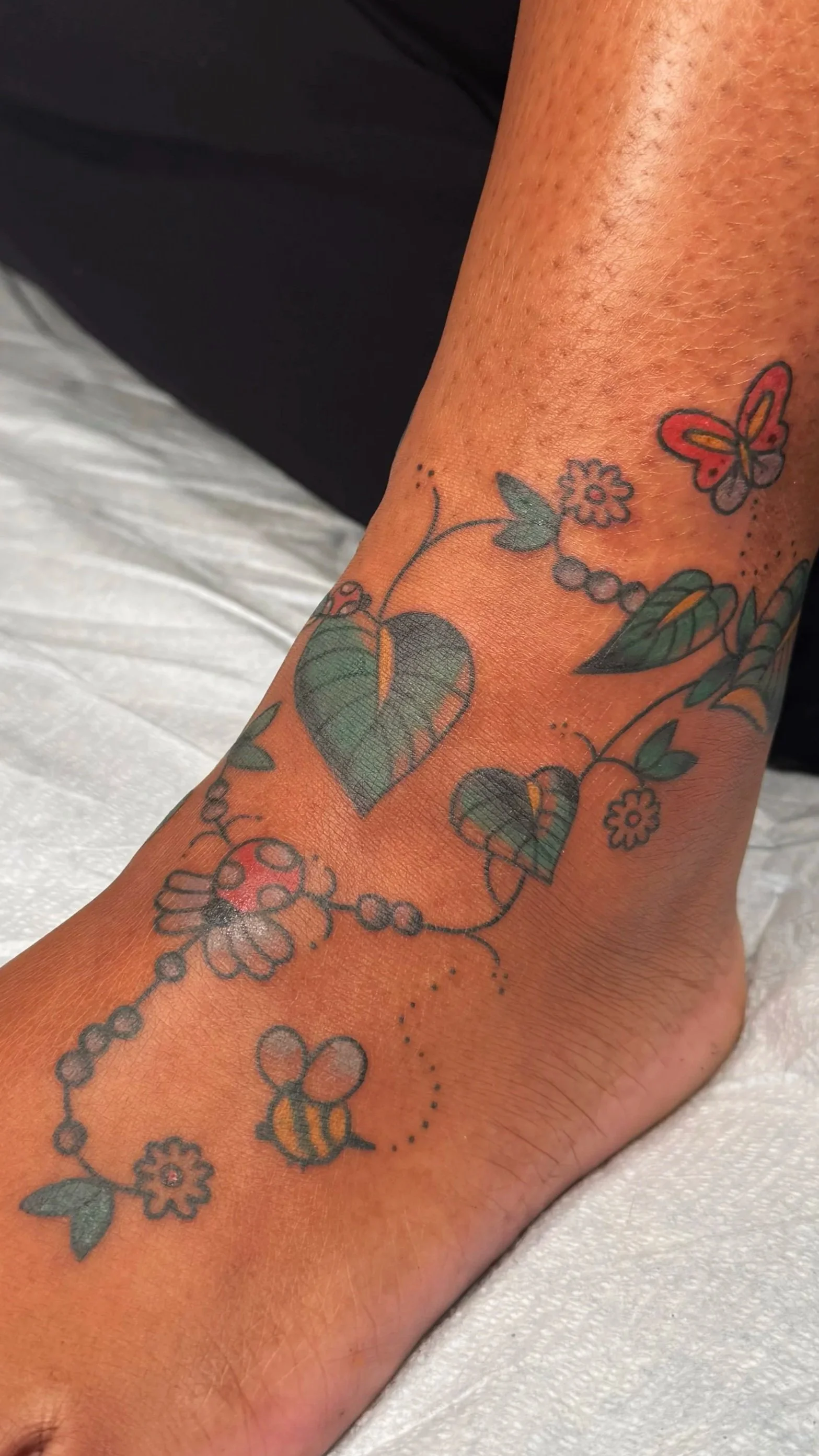 Tattoo of a butterfly, flowers, heart-shaped leaves, and a bee on a person's ankle.
