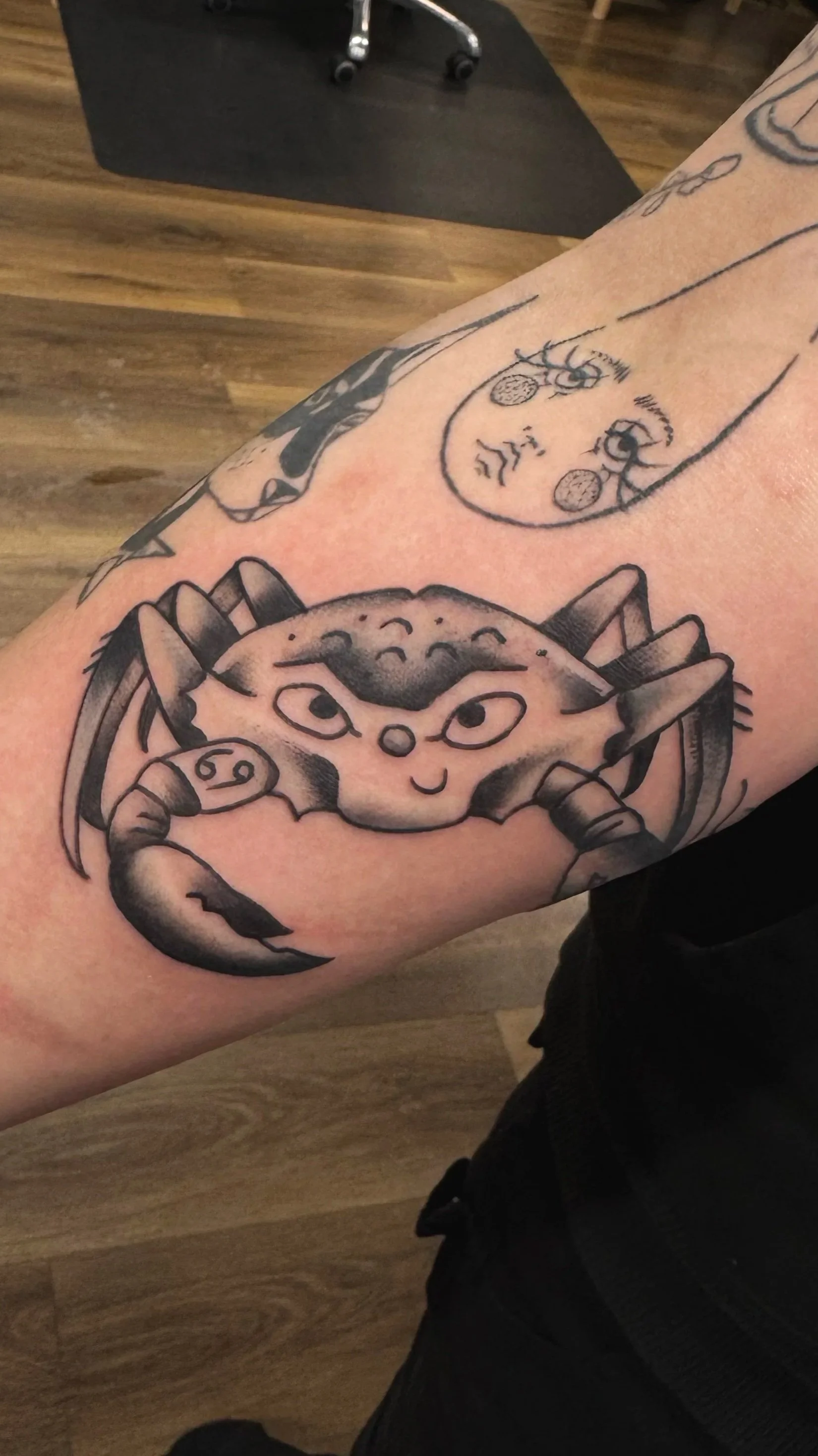 Tattoo of a cute cartoon crab with a smiling face on a person's arm, with some other tattoos visible nearby.