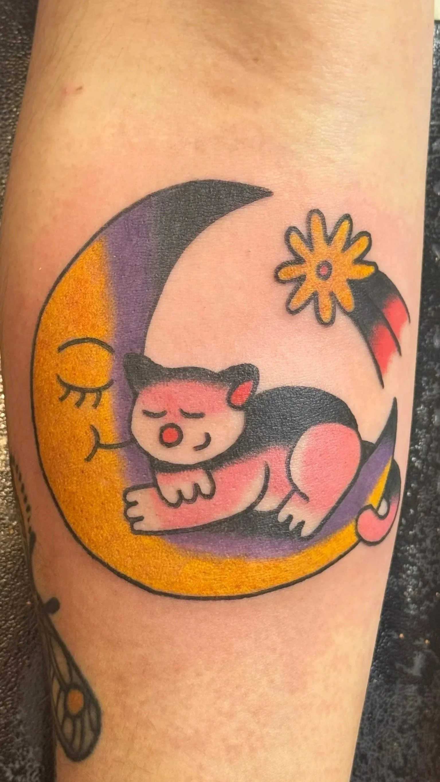 A tattoo of a sleeping black and white cat curled on a yellow crescent moon with a purple background, a smiling crescent moon face, a yellow shooting star with a red center, and a pink heart-shaped star.