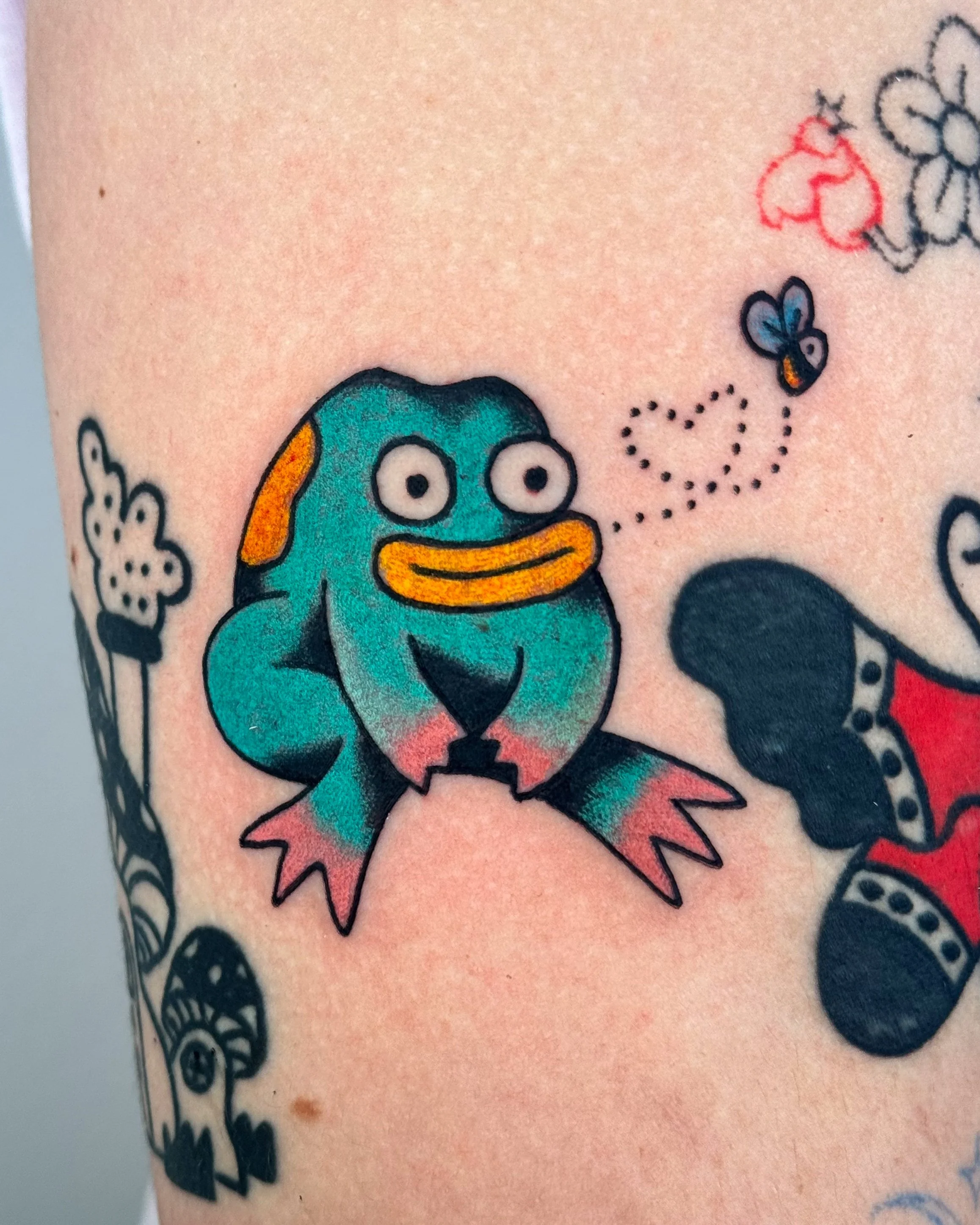 Colorful tattoo of a cartoon frog with big eyes and a wide smile, surrounded by other tattoos including a butterfly and hearts.