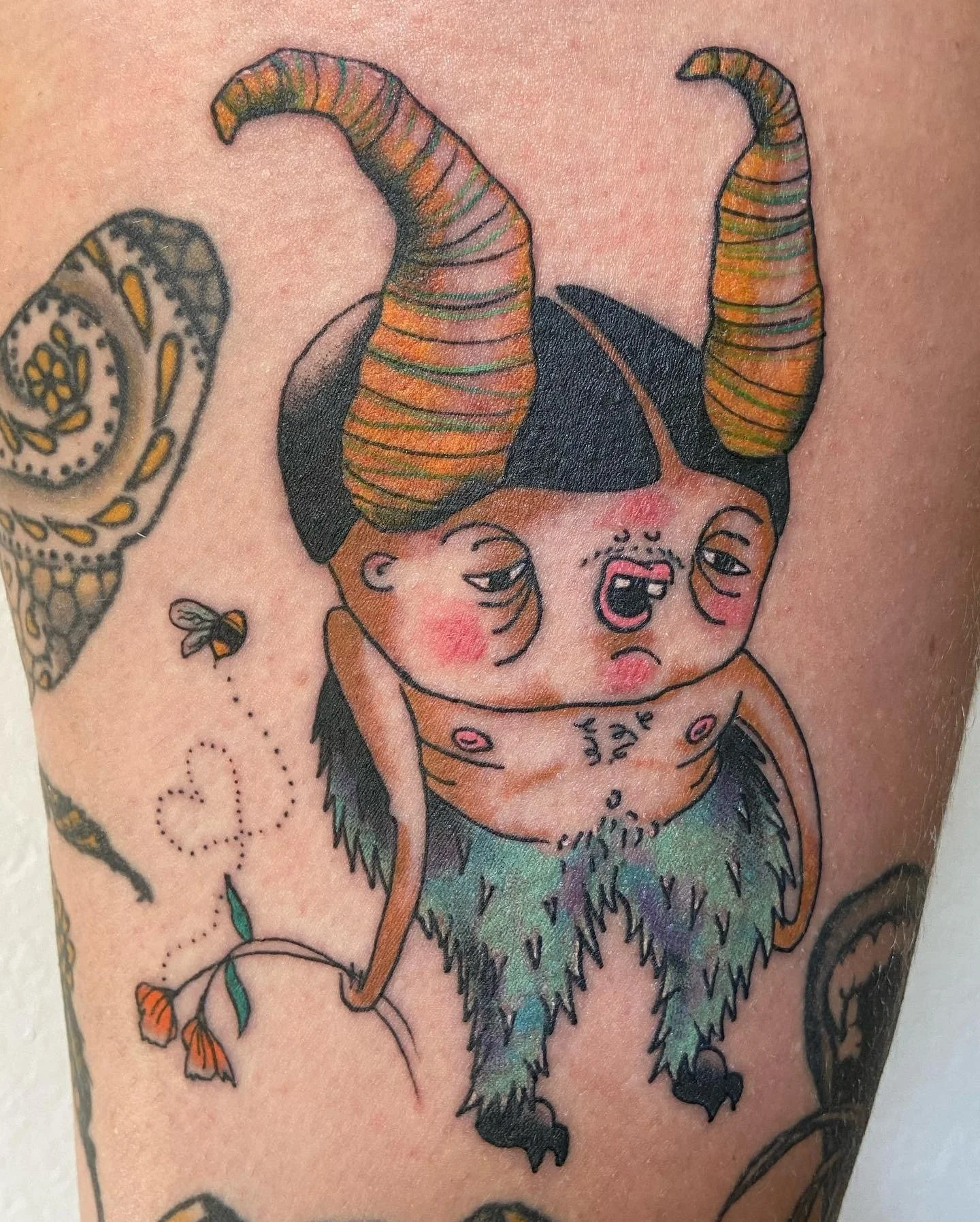 Tattoo of a creature with a human-like face and body, large twisted horns, and fuzzy colorful fur on the lower body, with other tattoo elements around it.