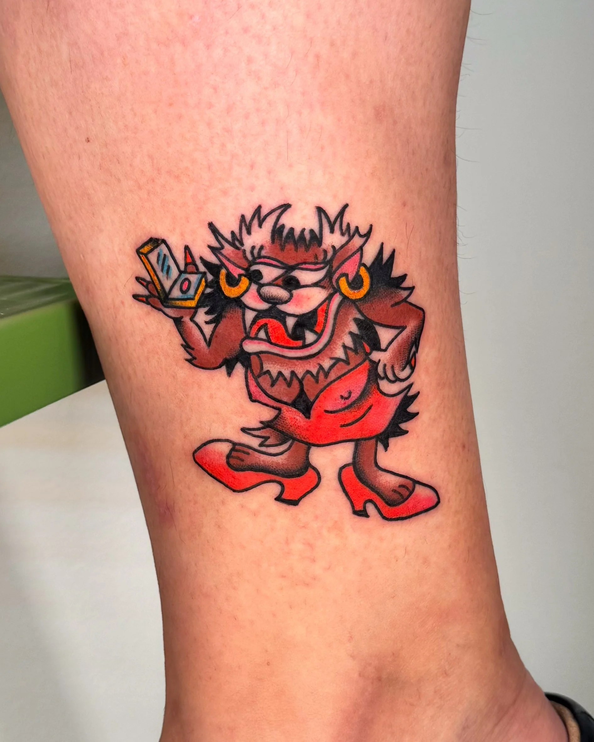Colorful tattoo of Taz with sunglasses, gold hoop earrings, holding a flip phone, wearing a red dress, red shoes, and standing on two legs.