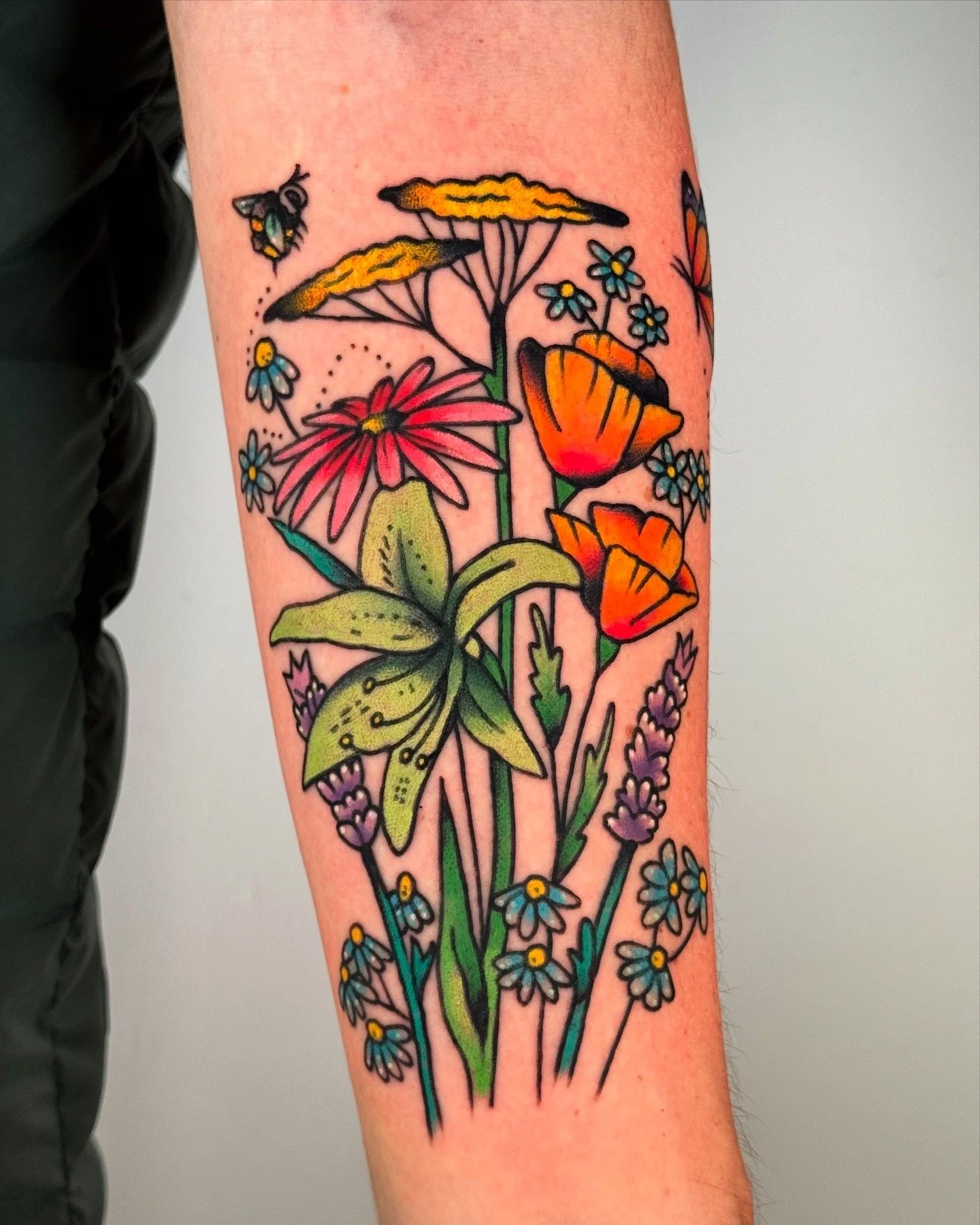 Colorful floral tattoo on forearm featuring daisies, lilies, poppies, grasses, and a bee.