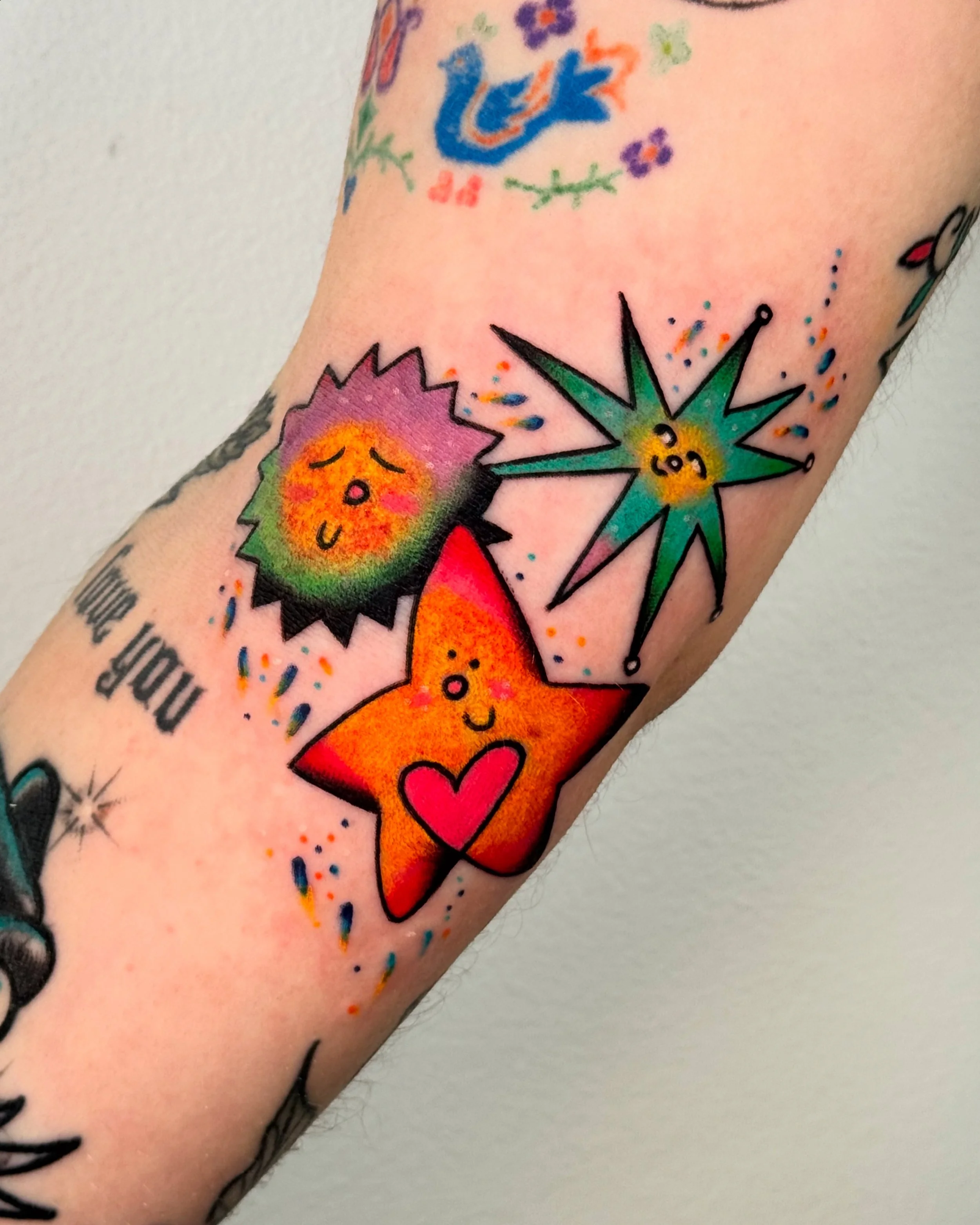 Colorful tattoo of three happy cartoon stars with faces, one orange with a pink heart, one green with a yellow center, and a black-spiked purple and green one, on a person's arm.