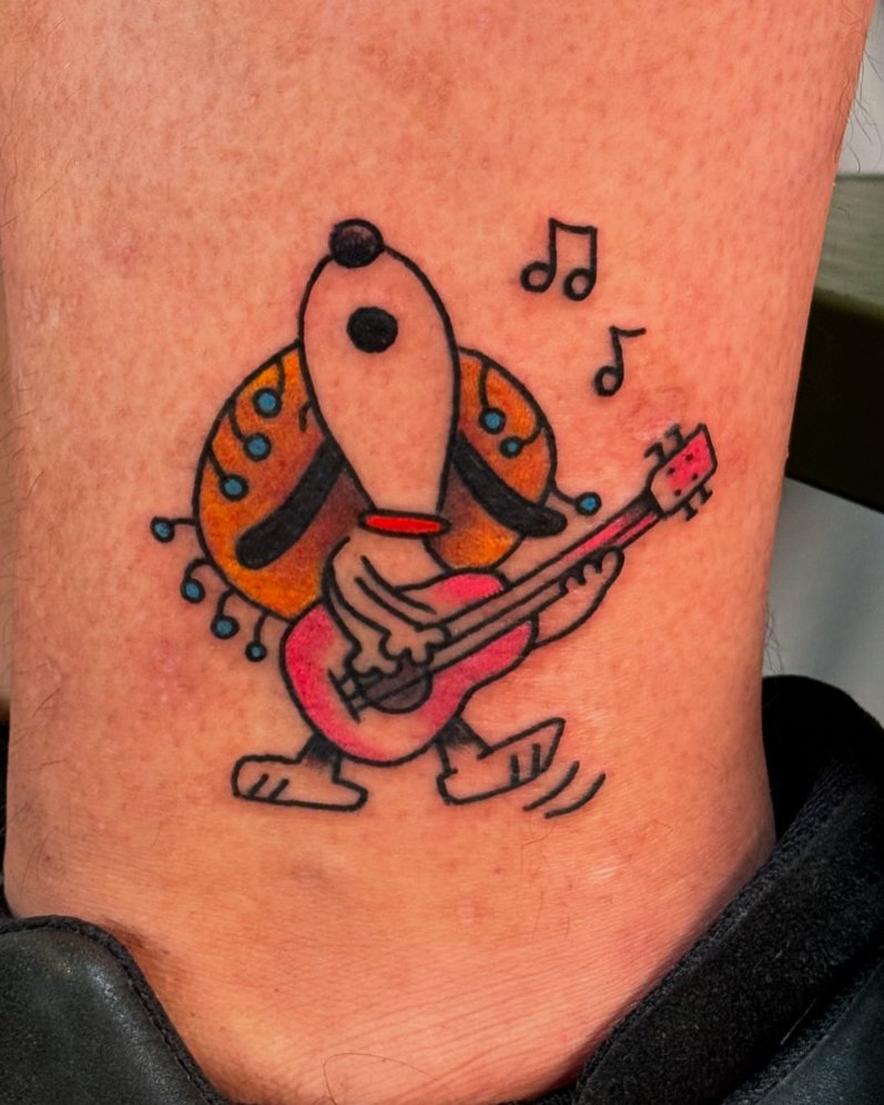 Tattoo of Snoopy the dog playing an acoustic guitar, with musical notes around him, on skin.