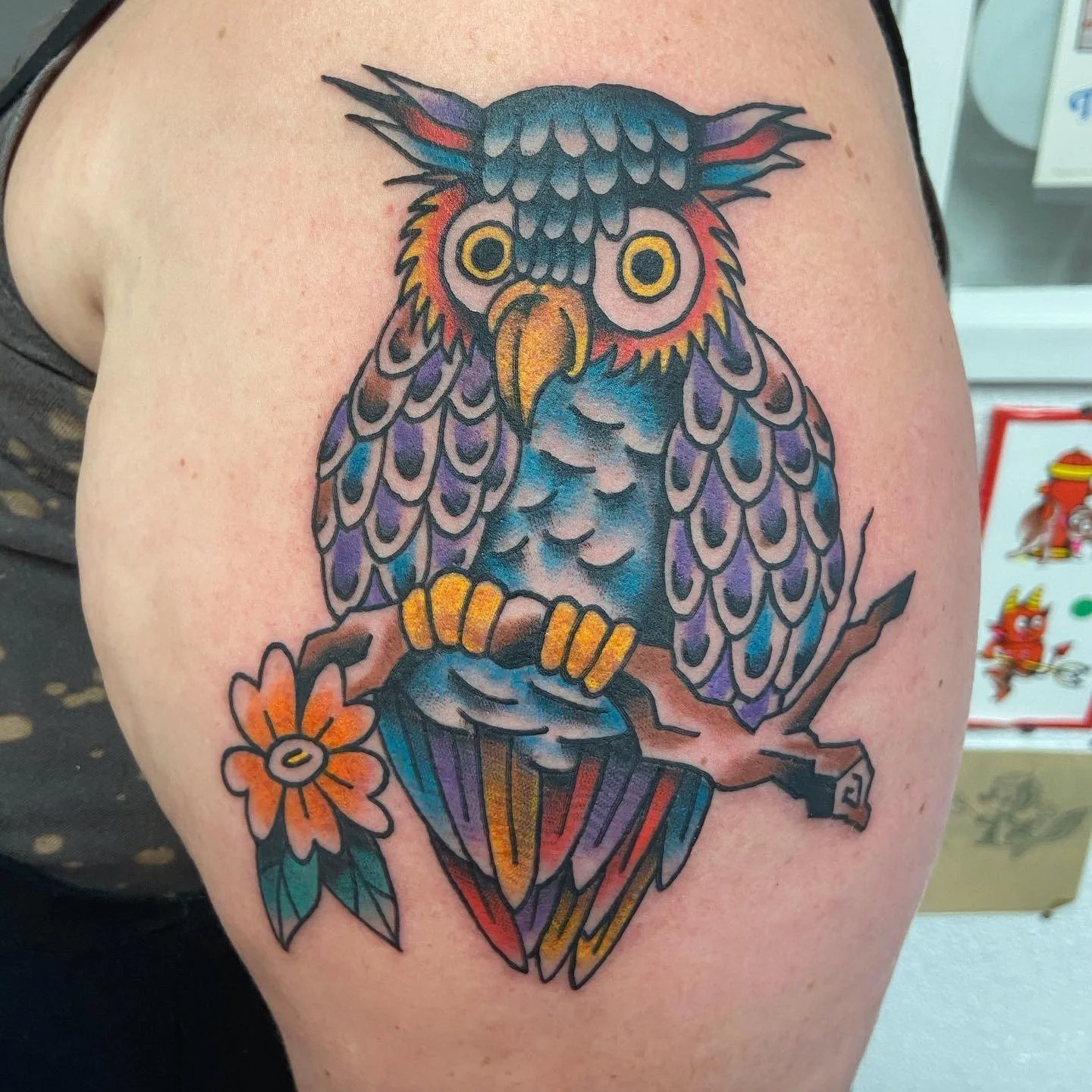 Colorful tattoo of an owl perched on a branch with a flower, featuring vibrant shades of blue, purple, yellow, and orange.