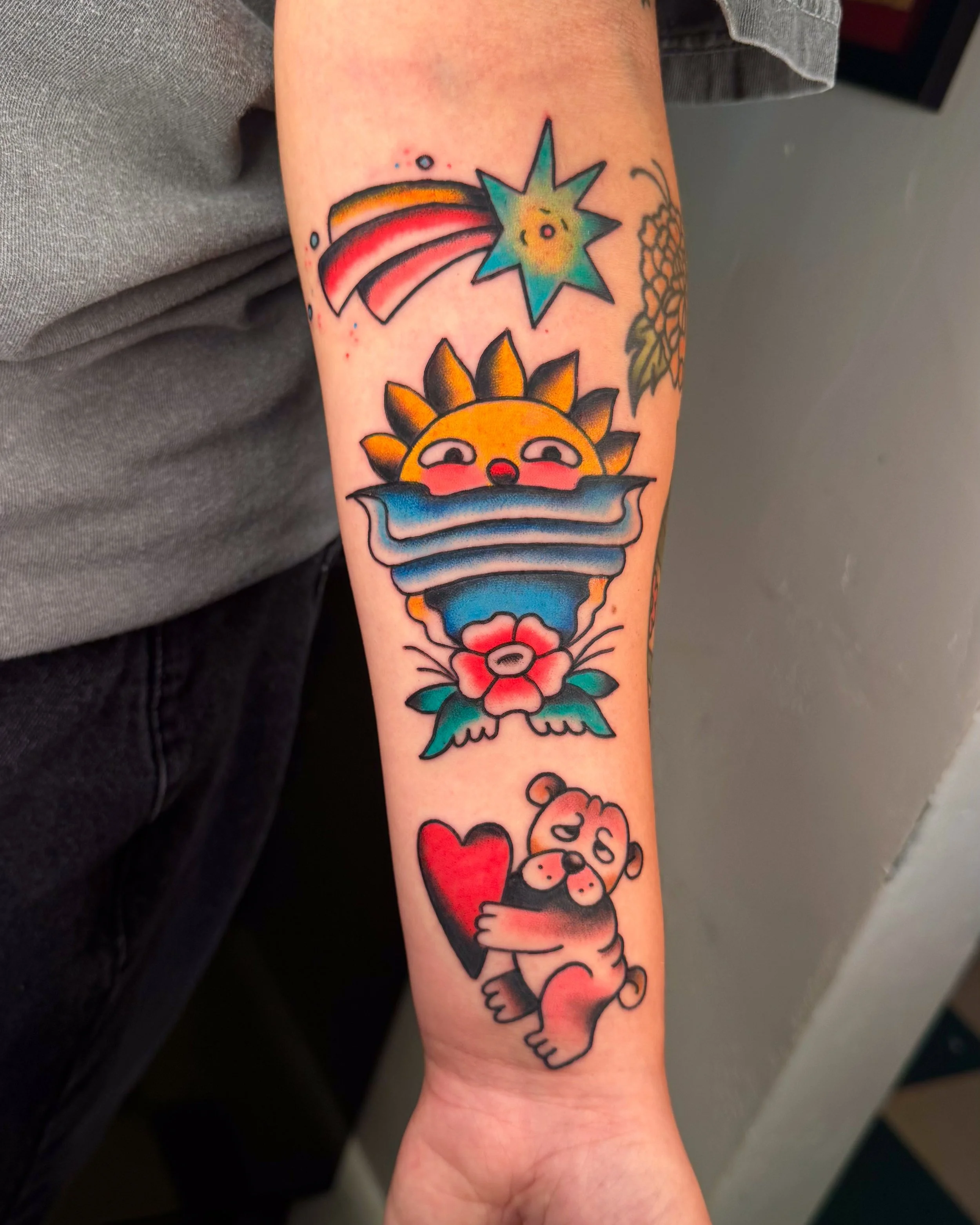 Colorful tattoo on arm featuring a smiling sun with half-closed eyes, blue waves, a red flower, a shooting star, and a cute bear hugging a red heart.
