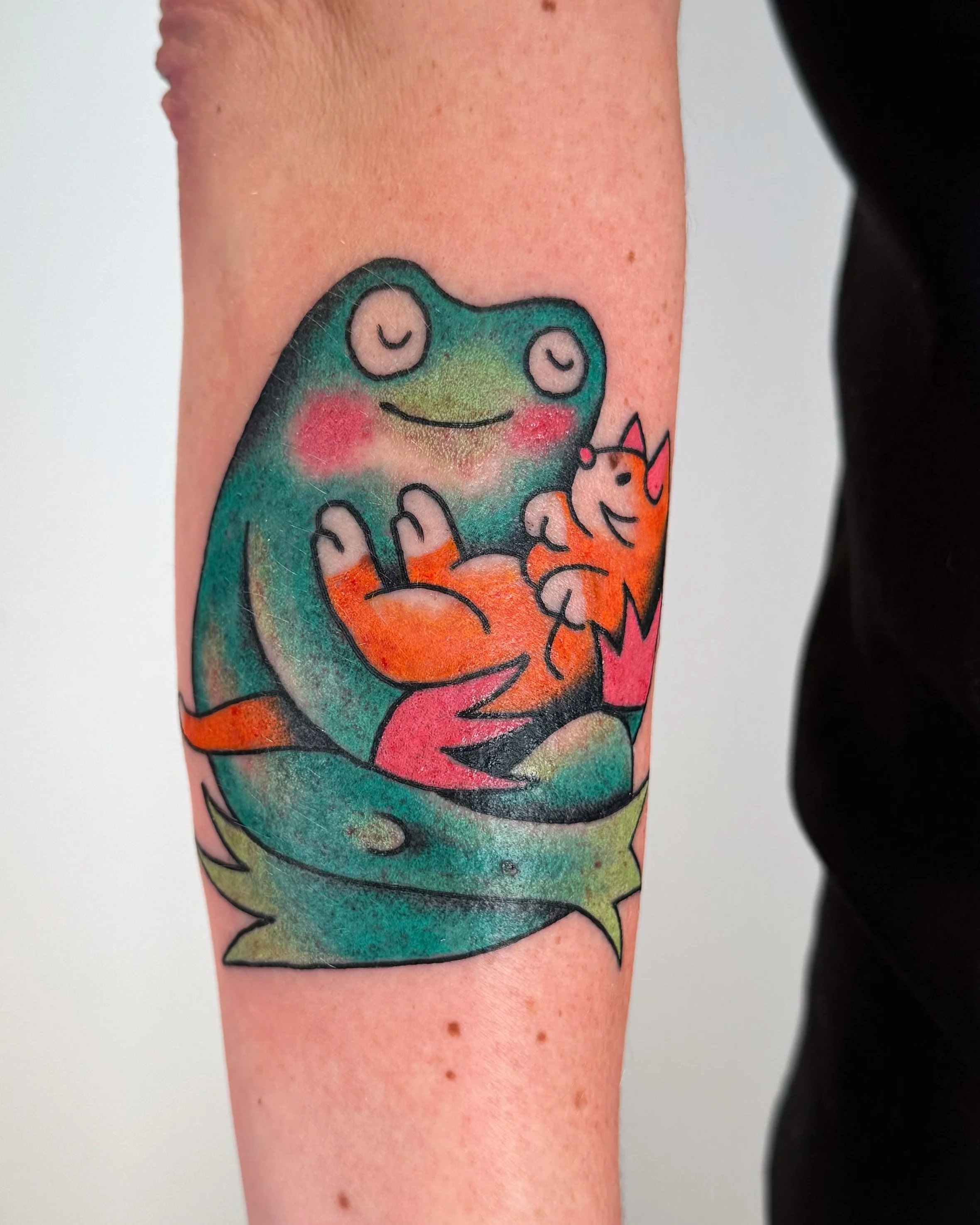 Colorful tattoo of a cartoon frog hugging a cartoon orange cat.