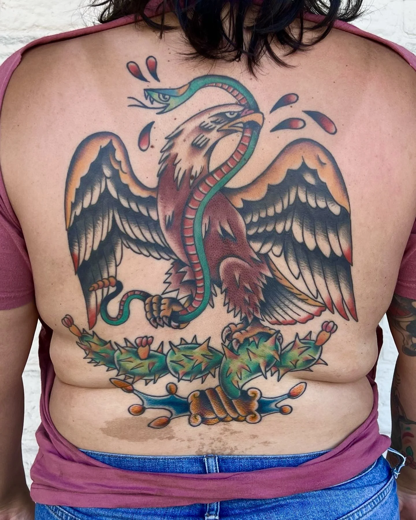 Colorful tattoo on person's back of a reimagined design from the Mexican flag: an eagle with outstretched wings, a snake intertwined with its body, and a banner with laurel leaves at the bottom.