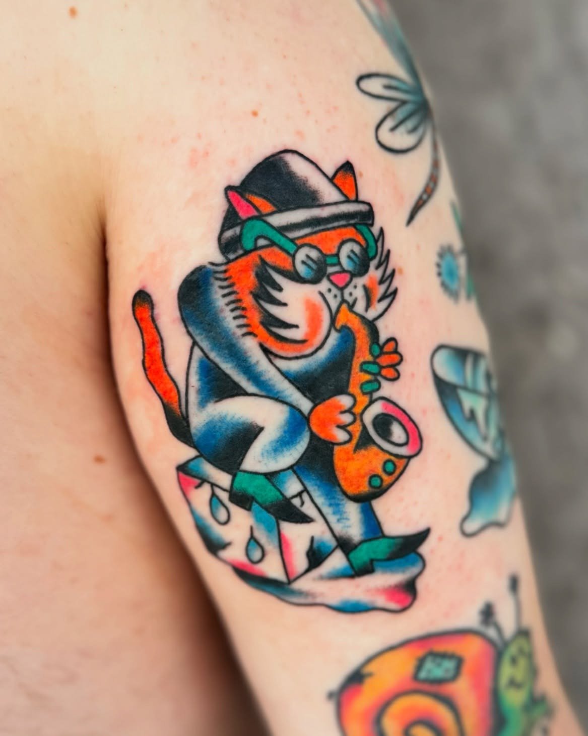 Colorful tattoo of a cat playing saxophone, wearing sunglasses and a hat, with traditional tattoo style.