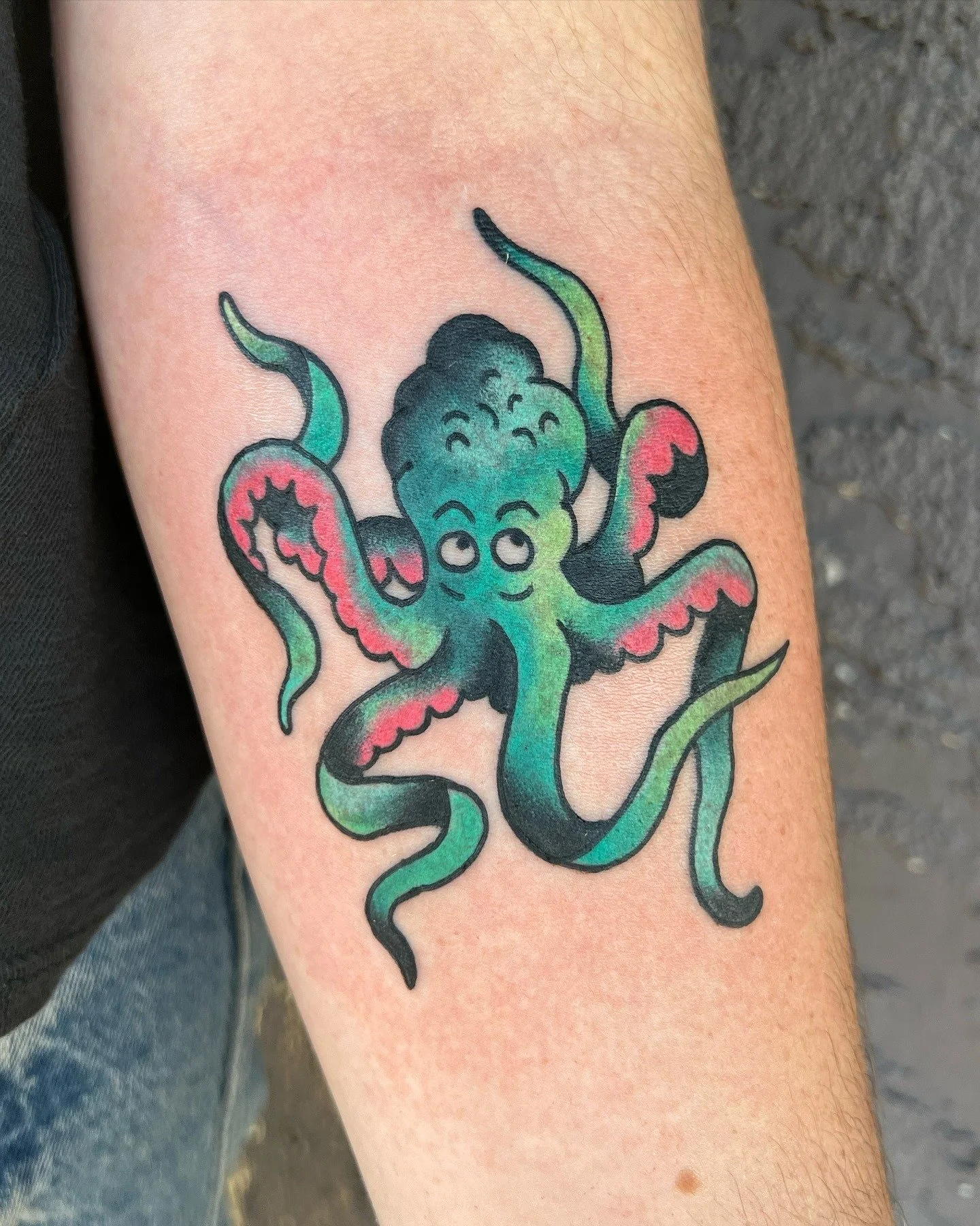 Colorful tattoo of a cute, cartoon-style octopus on a person's arm, with pink, green, and black ink details.