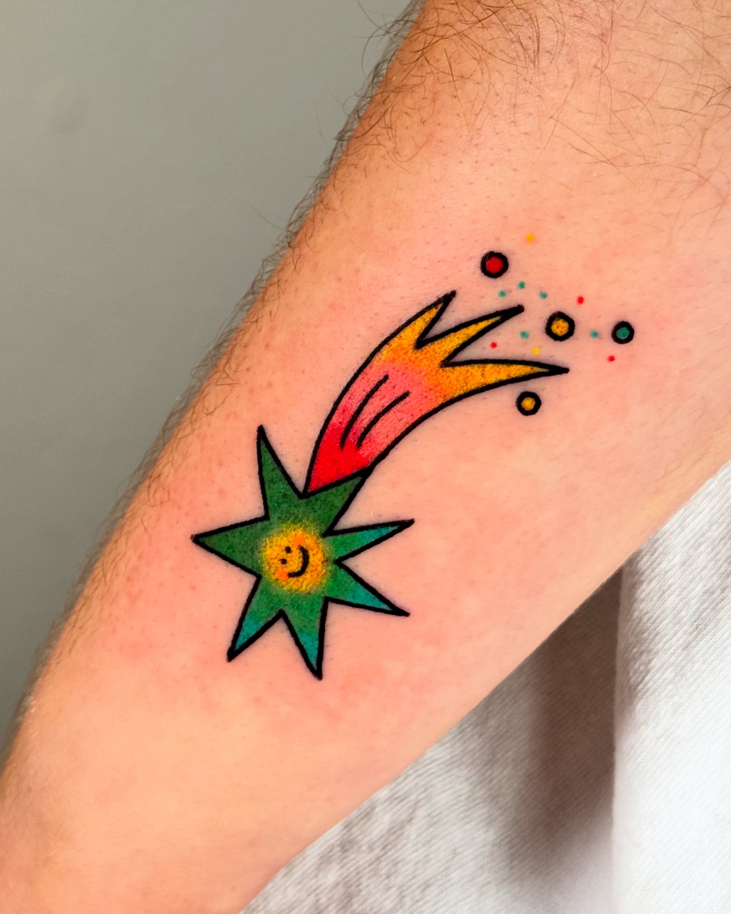 Colorful tattoo of a shooting star with a smiley face, shotting stars and dots, on person's arm.