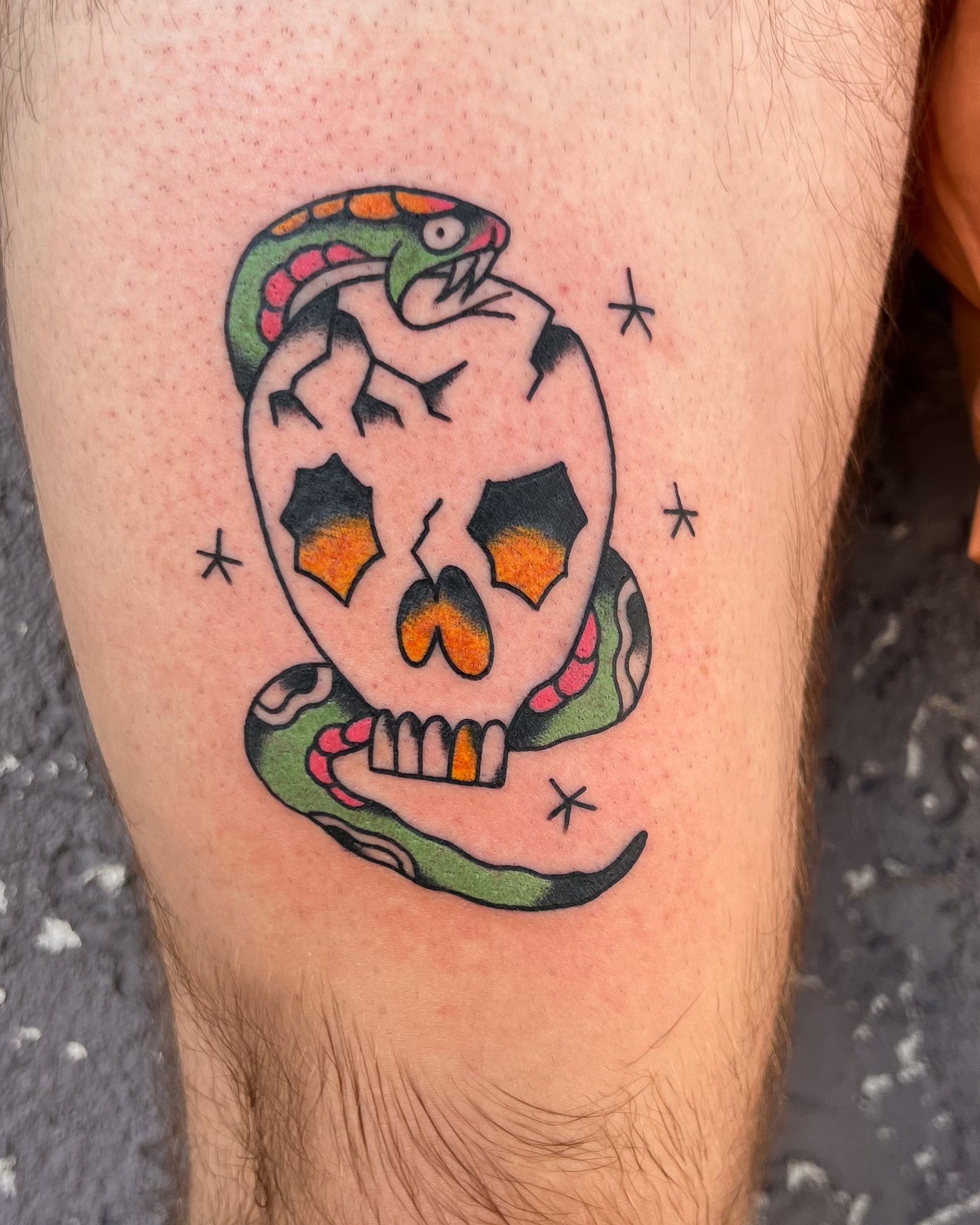 Tattoo of a skull with orange and black eye sockets and nose, a pink and green snake wrapped around it, and a colorful snake-like creature on top of the skull, with small black stars around.