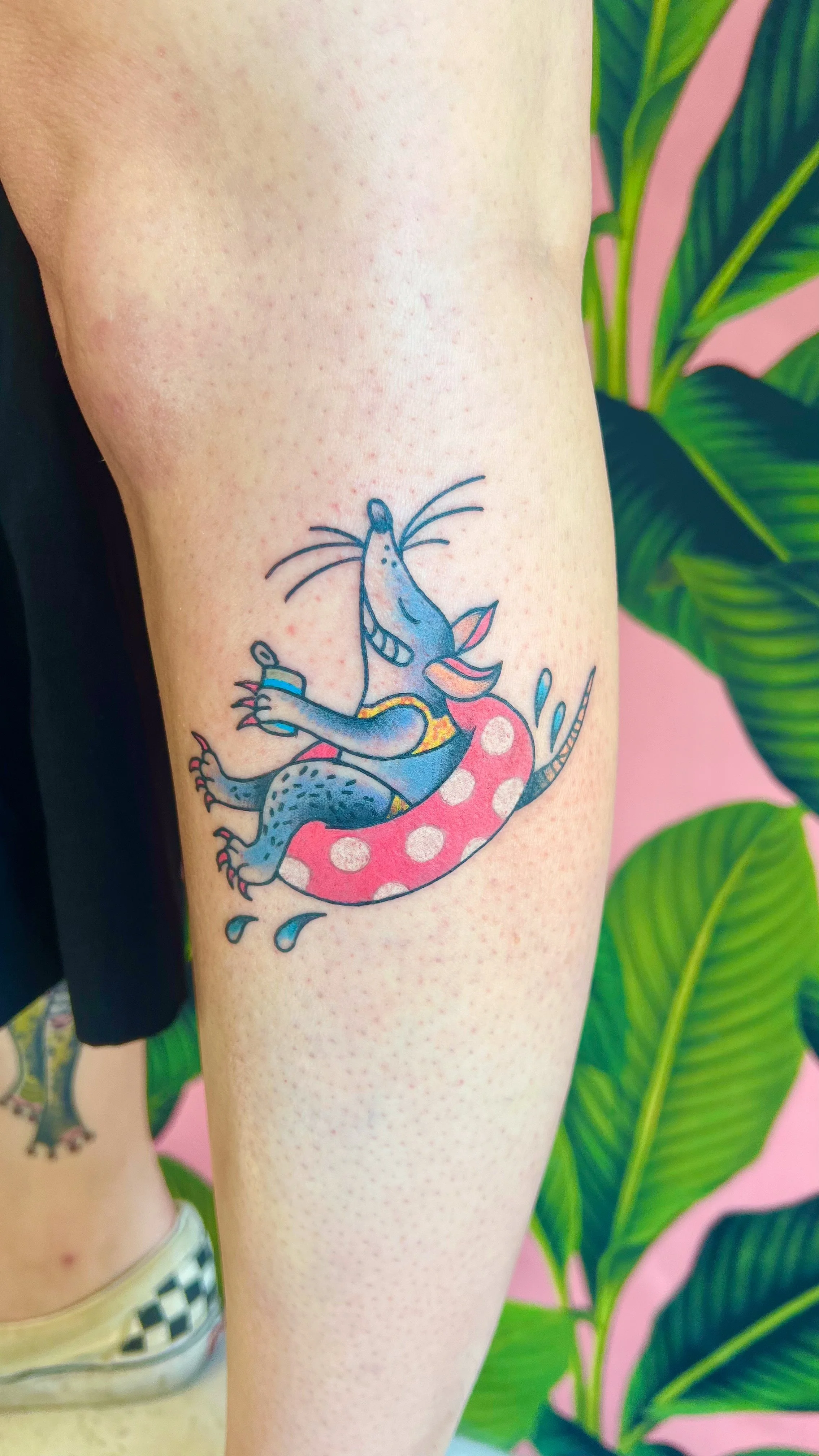 Colorful tattoo of a Blue Rat with a yellow shirt riding on a pink polka dot pool float, smiling and holding a drink.