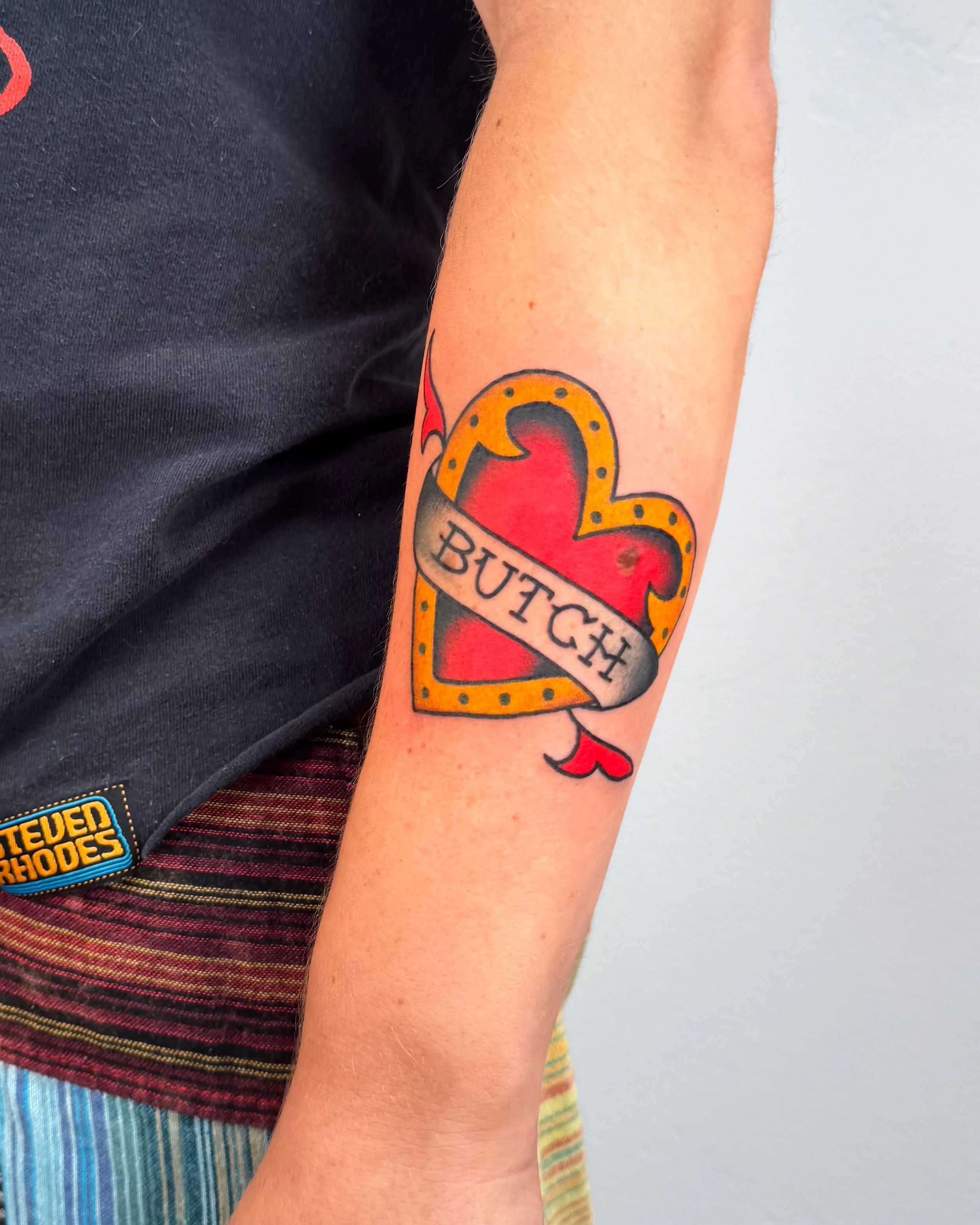 Tattoo of a red heart inside a yellow-outlined heart shape with small dots, with a grey banner across it bearing the word 'BUTCH' in black capital letters.
