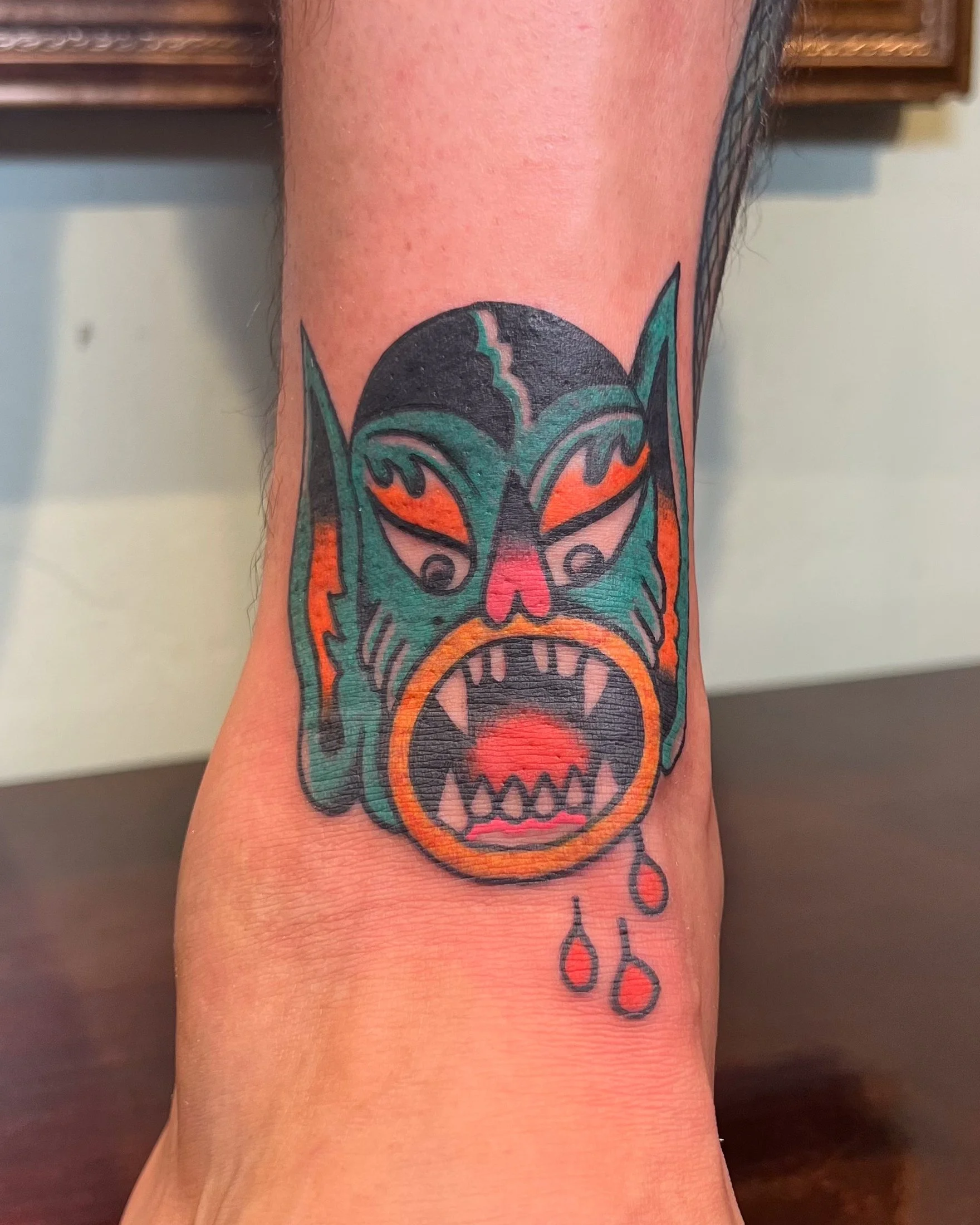 Colorful tattoo of a fierce, menacing creature with sharp teeth, pointed ears, and a glowing mouth, featuring dripping blood elements.