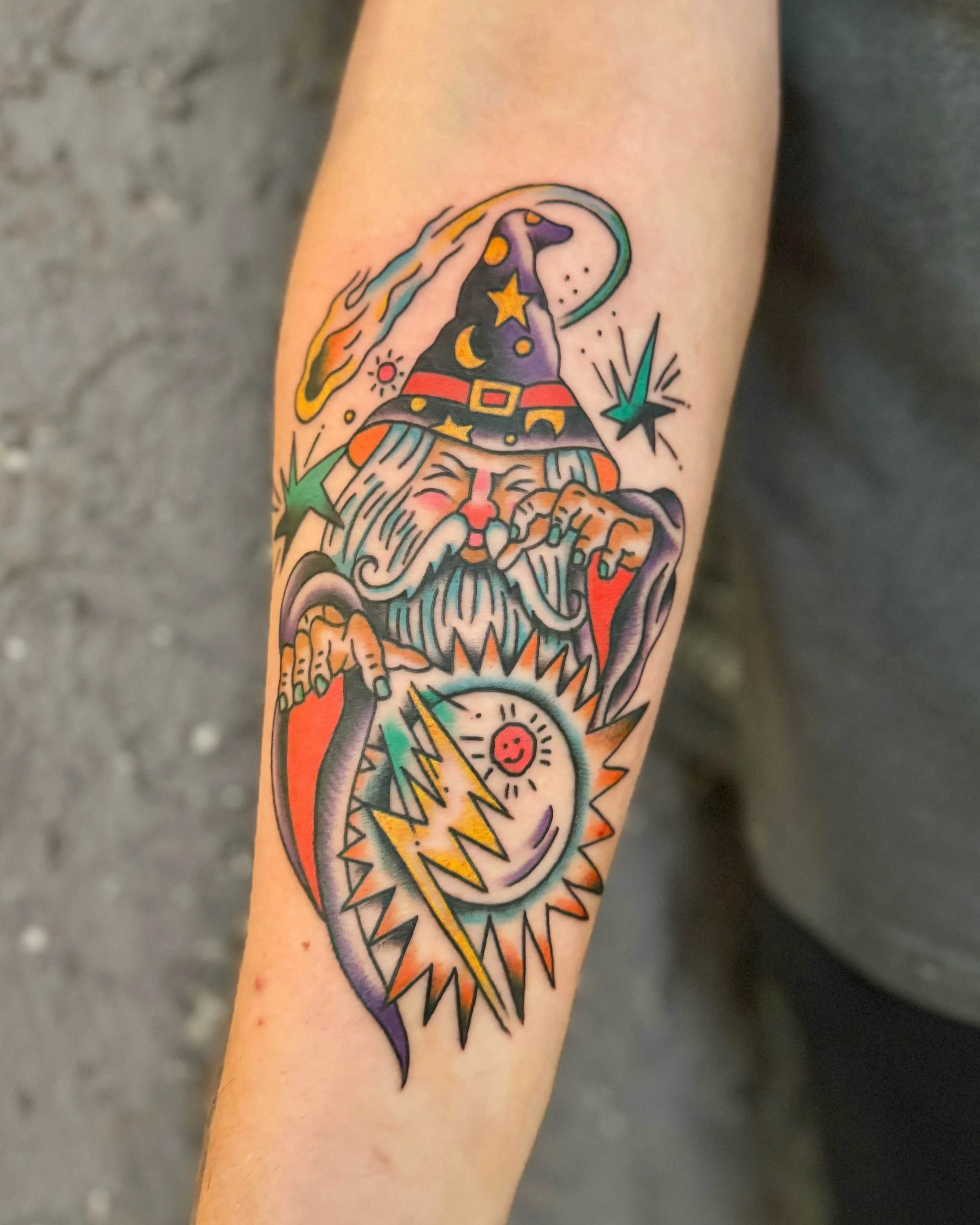 A colorful tattoo of a wizard with a long beard wearing a starry hat and robe, holding a magic staff, surrounded by stars and abstract designs.