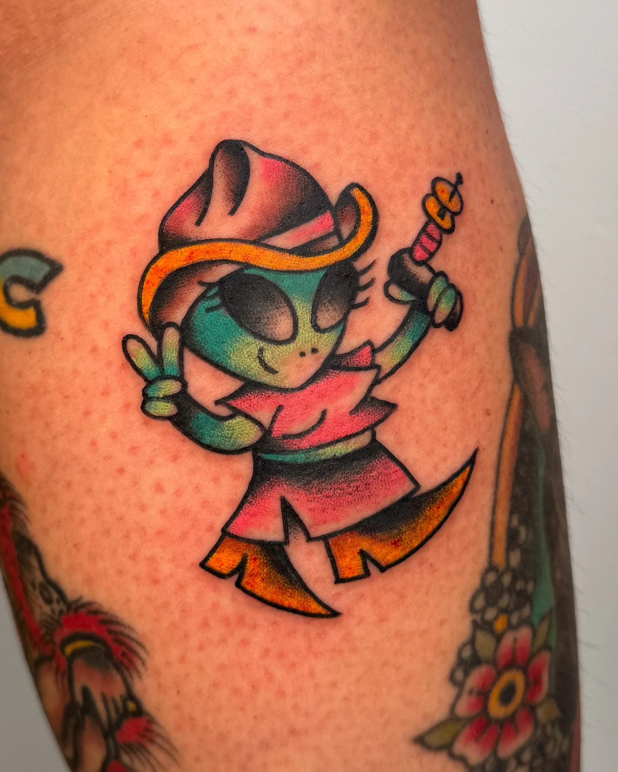 A colorful tattoo of a cartoon alien dressed as a cowgirl, wearing a hat and holding a toy sword, with hand signal and a smile on its face.