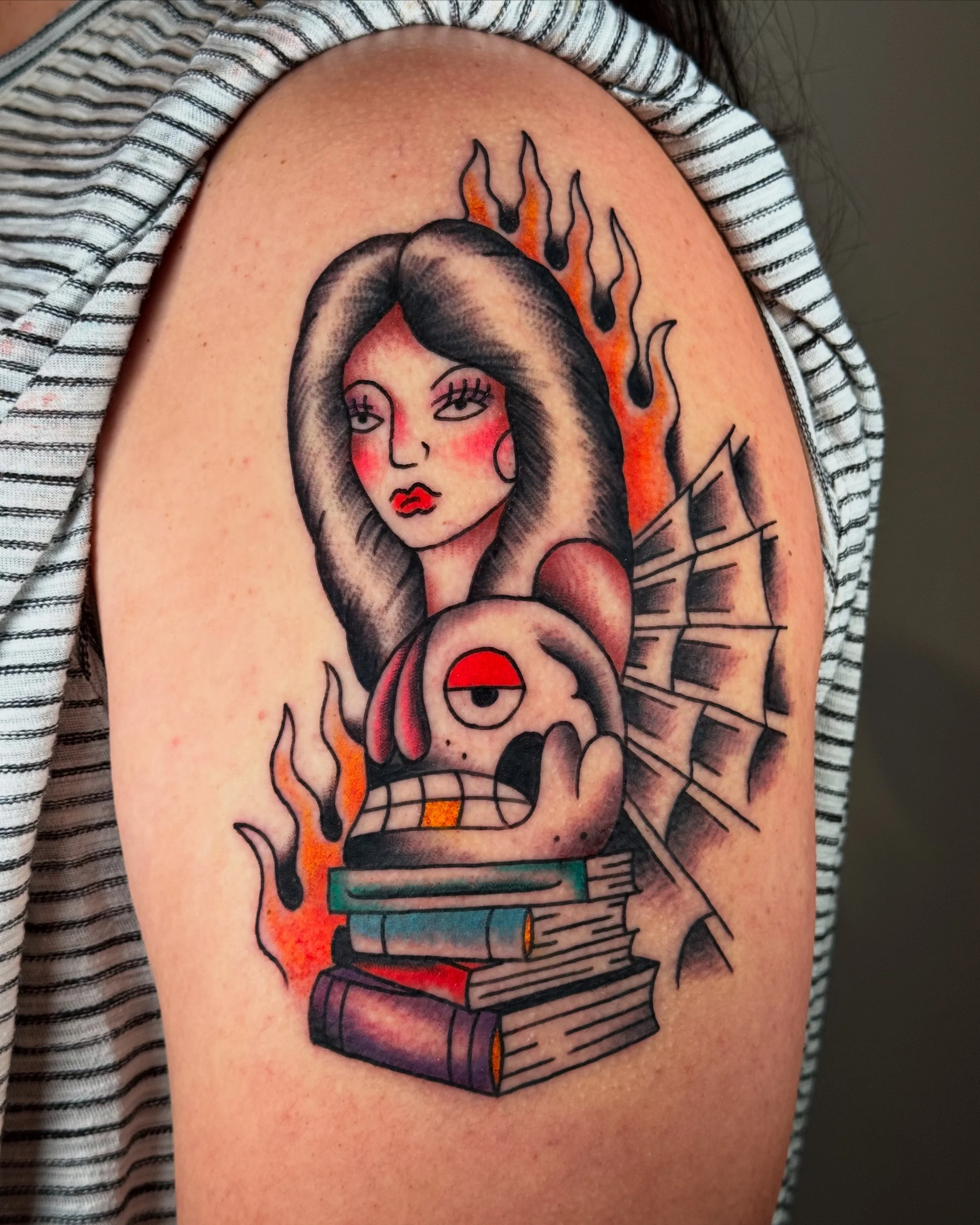 Tattoo of a woman with dark hair and red blush, a cartoonish creature with one eye, books, flames, and a newspaper background on upper arm.
