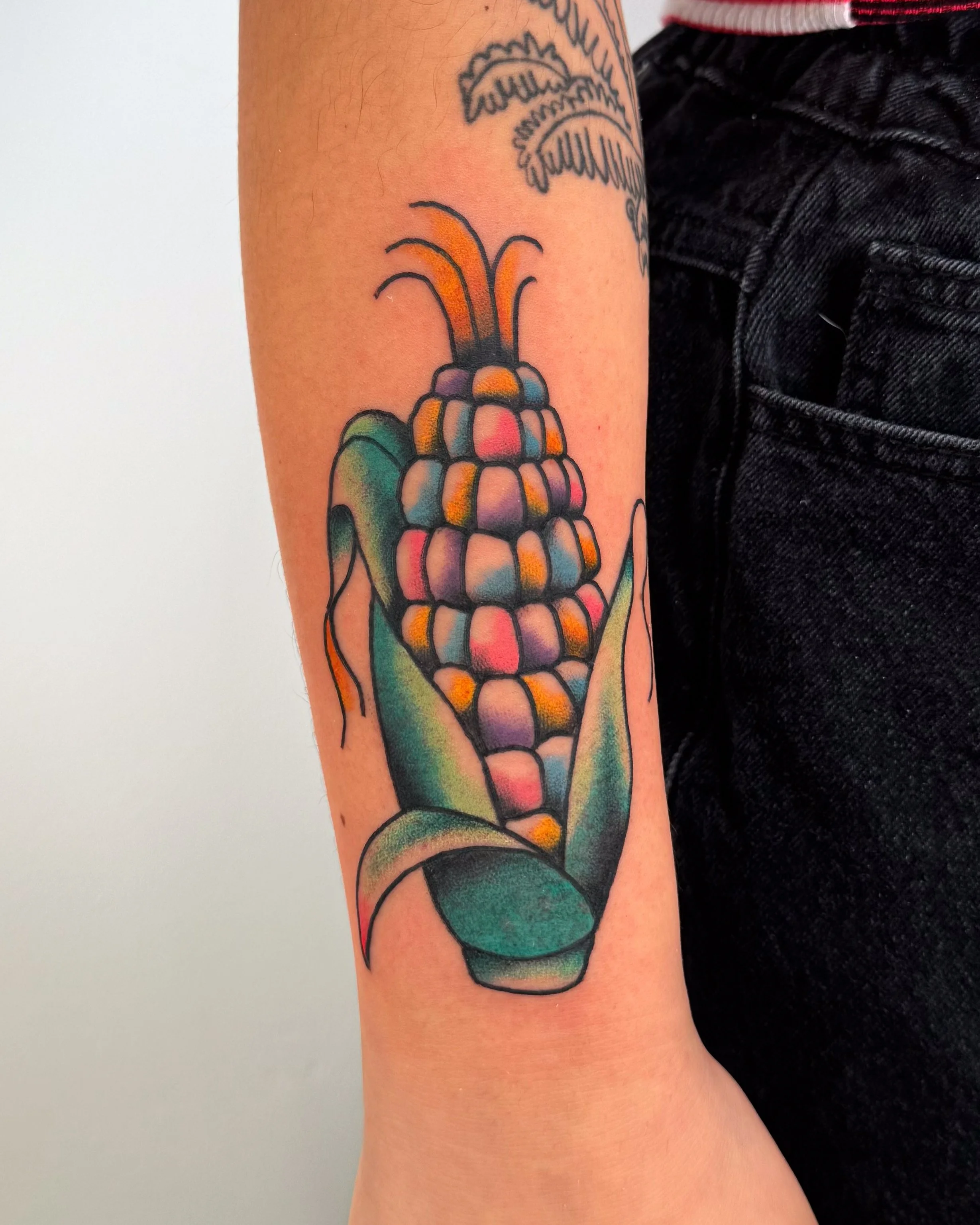 Colorful tattoo of an ear of corn with green leaves on a person's arm.