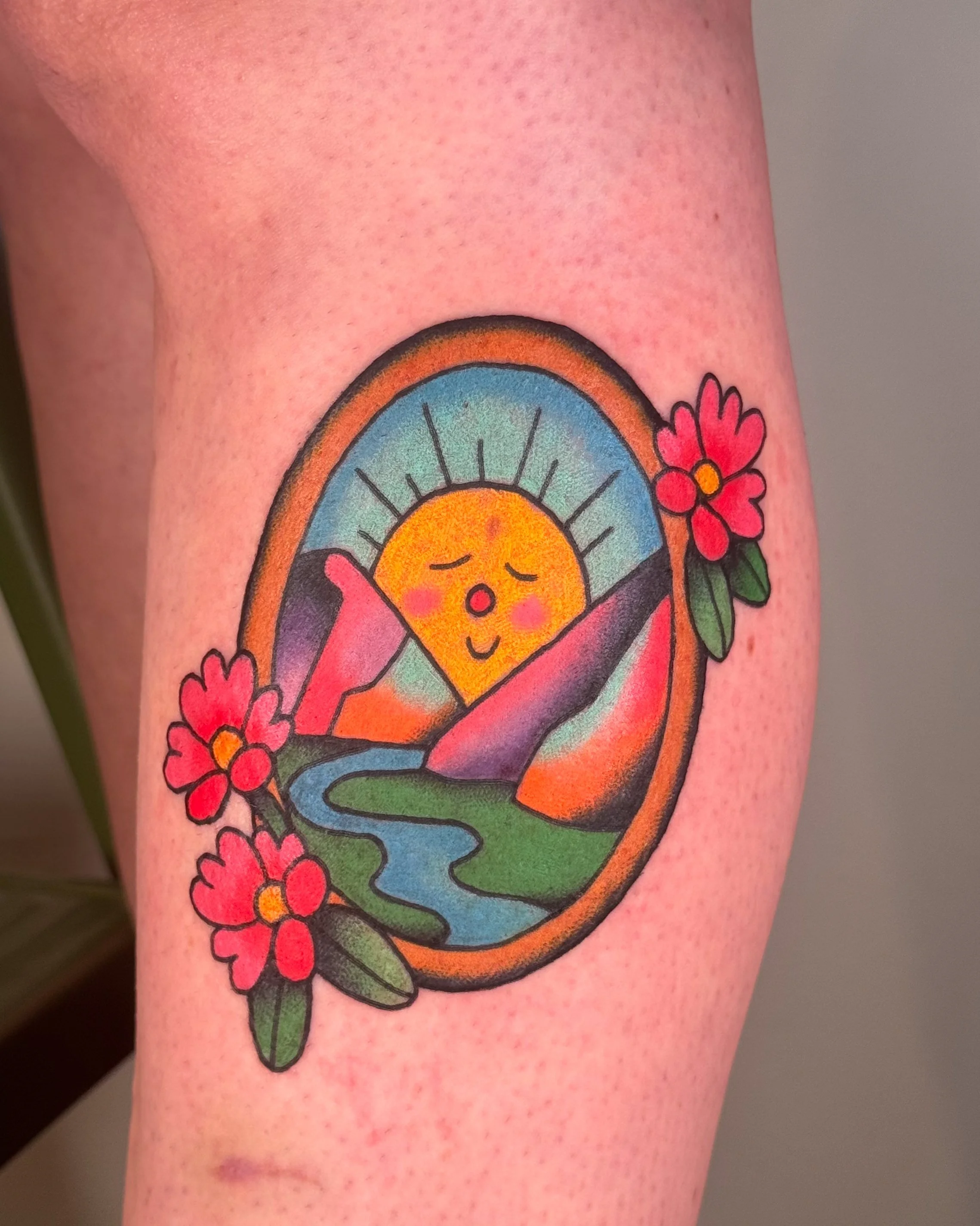 Colorful tattoo of a smiling sun with closed eyes, surrounded by mountains, a river, a rainbow, and pink flowers on a person's skin.