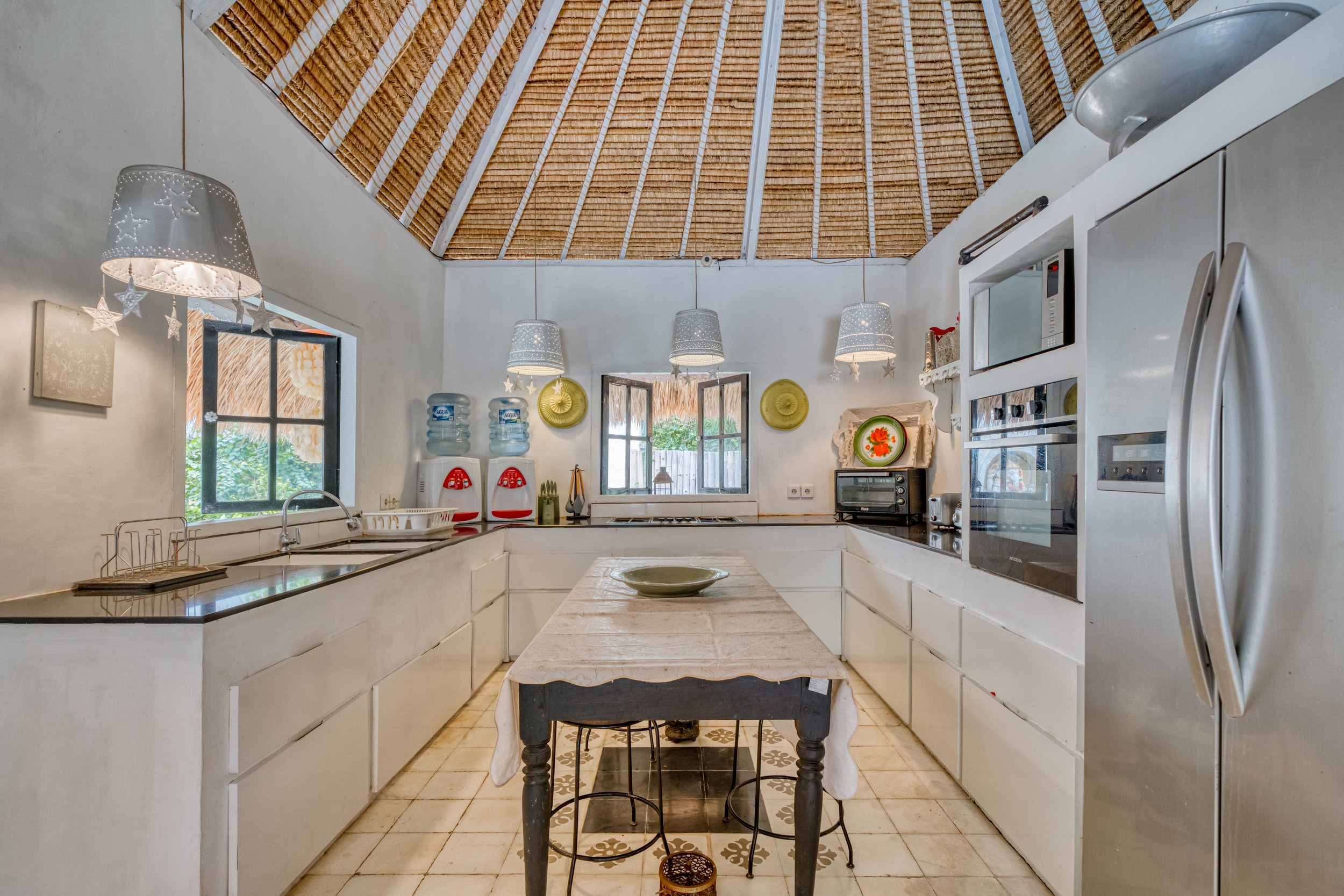 A bright, rustic kitchen with a thatched roof, white cabinets, and small window. Features include a microwave, oven, water dispensers, and a small dining table with a bowl. Hanging lamps with decorative shades, plates on the wall, and outdoor scenery visible through the window.