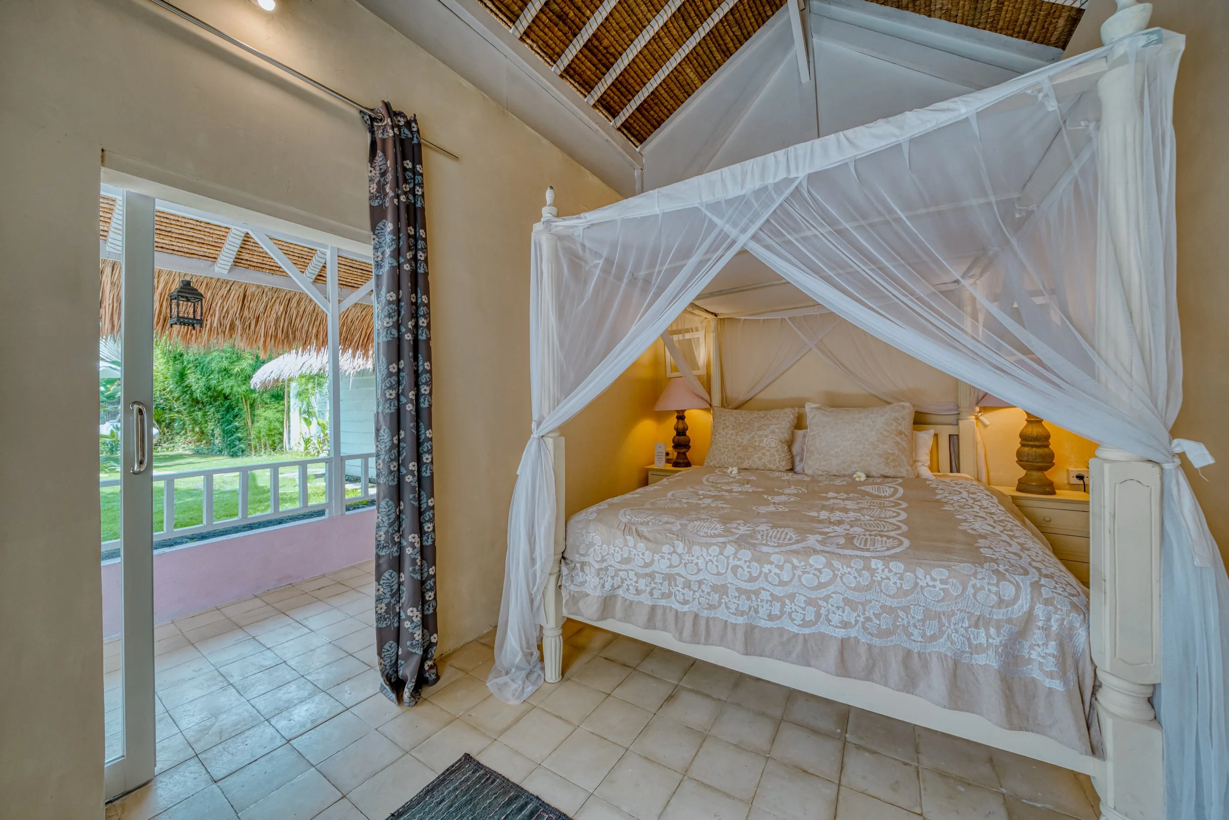 Indoor bedroom with a four-poster bed canopy, beige linens, and a nightstand with a lamp. An open door leads to a balcony with greenery and thatched roof structures outside.