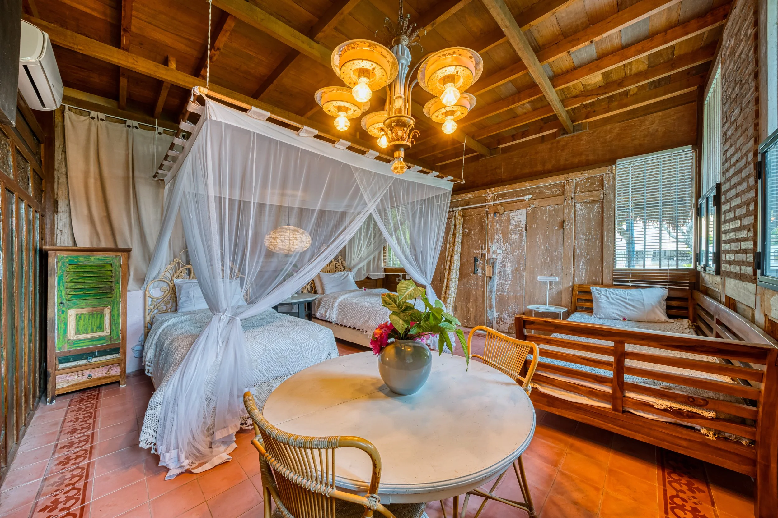 A rustic bedroom with wooden walls and ceiling, including two beds with white bedding, a canopy with mosquito netting, a round dining table with a flower vase, wicker chairs, and a single daybed near the window. The room features a vintage chandelier, tiled floor, and natural light from the windows.