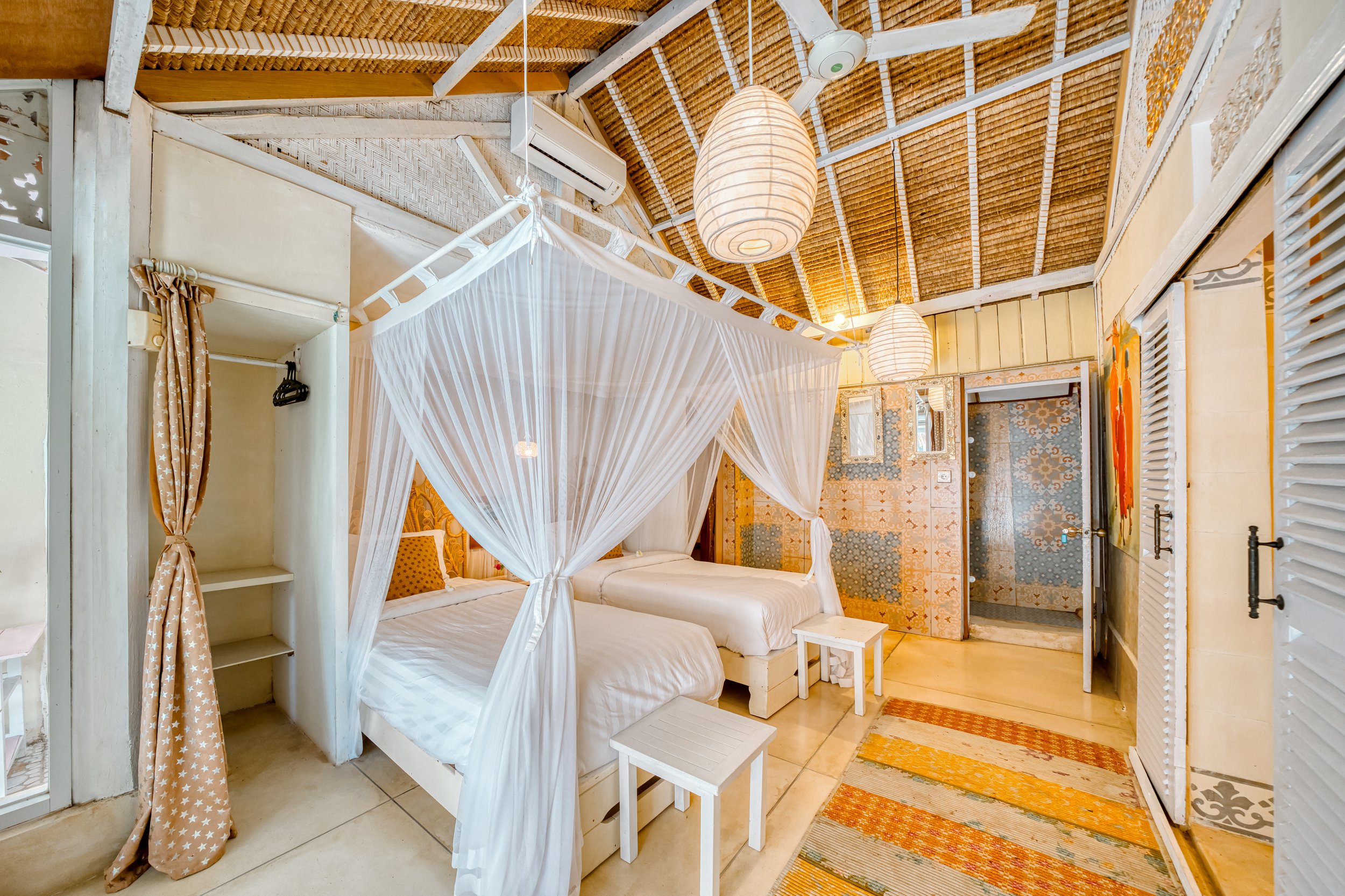 A cozy bedroom with two beds covered by white mosquito nets, warm lighting, wooden accents, patterned walls, and a colorful rug.