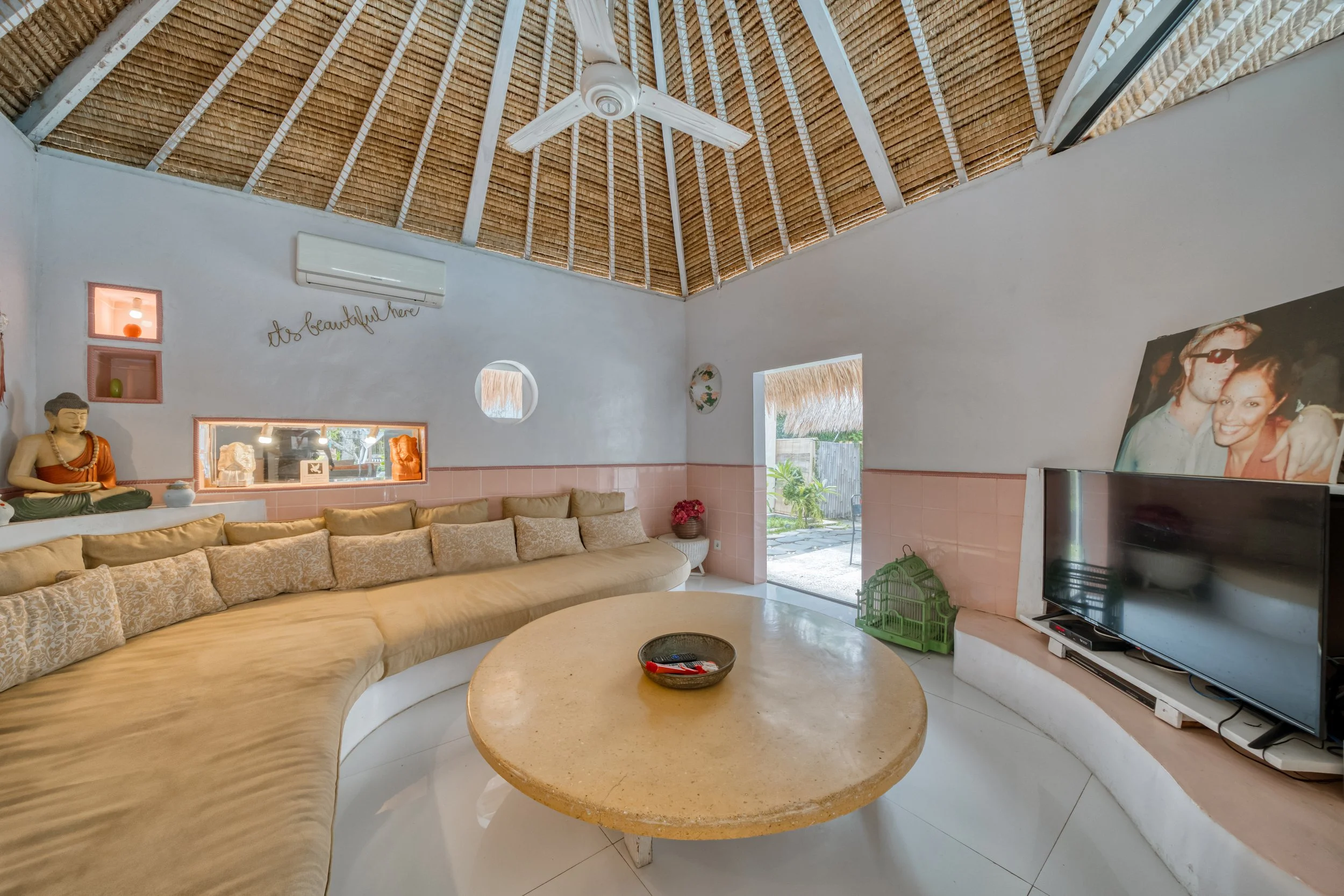 Living room with a curved beige sofa, a round coffee table, a large TV, decorative Buddha statue, wall art, and an open door revealing a garden outside.