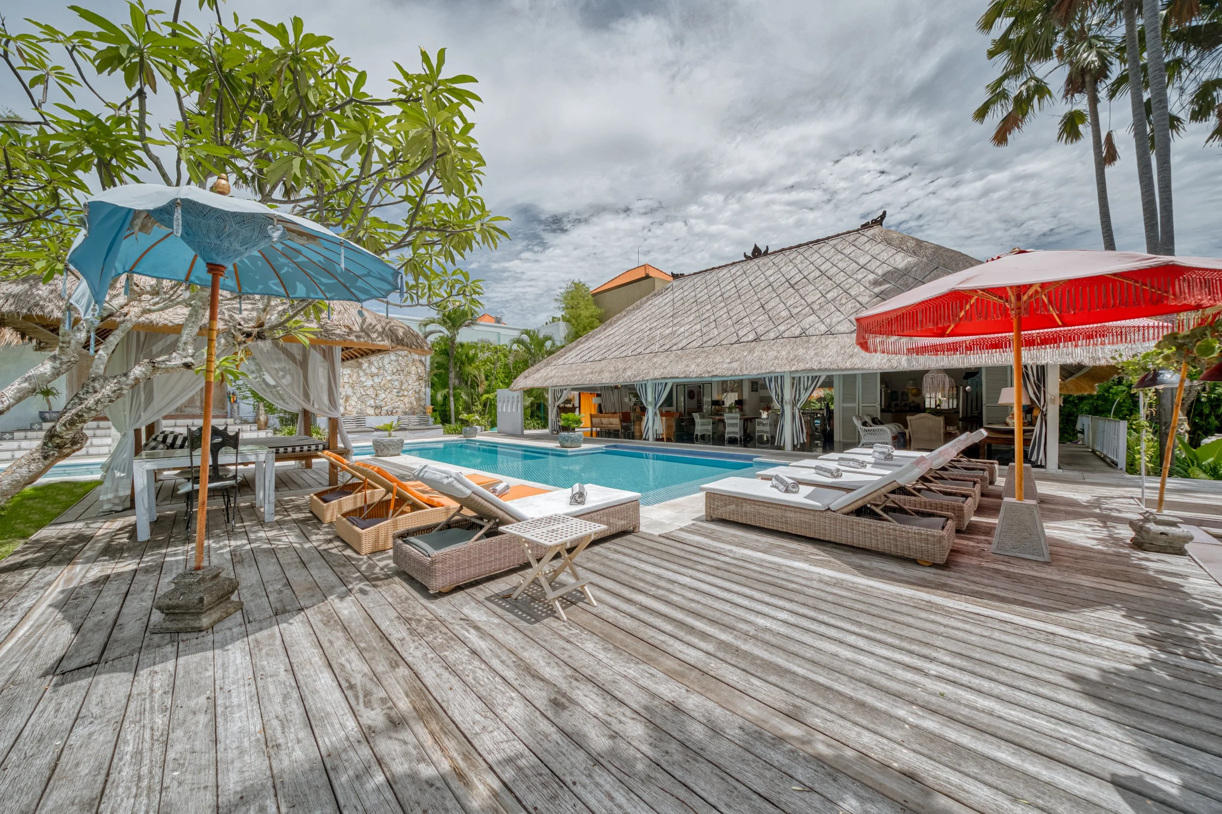 A luxurious outdoor pool area with wooden deck, lounge chairs, umbrellas, and a thatched-roof cabana, surrounded by greenery on a partly cloudy day.