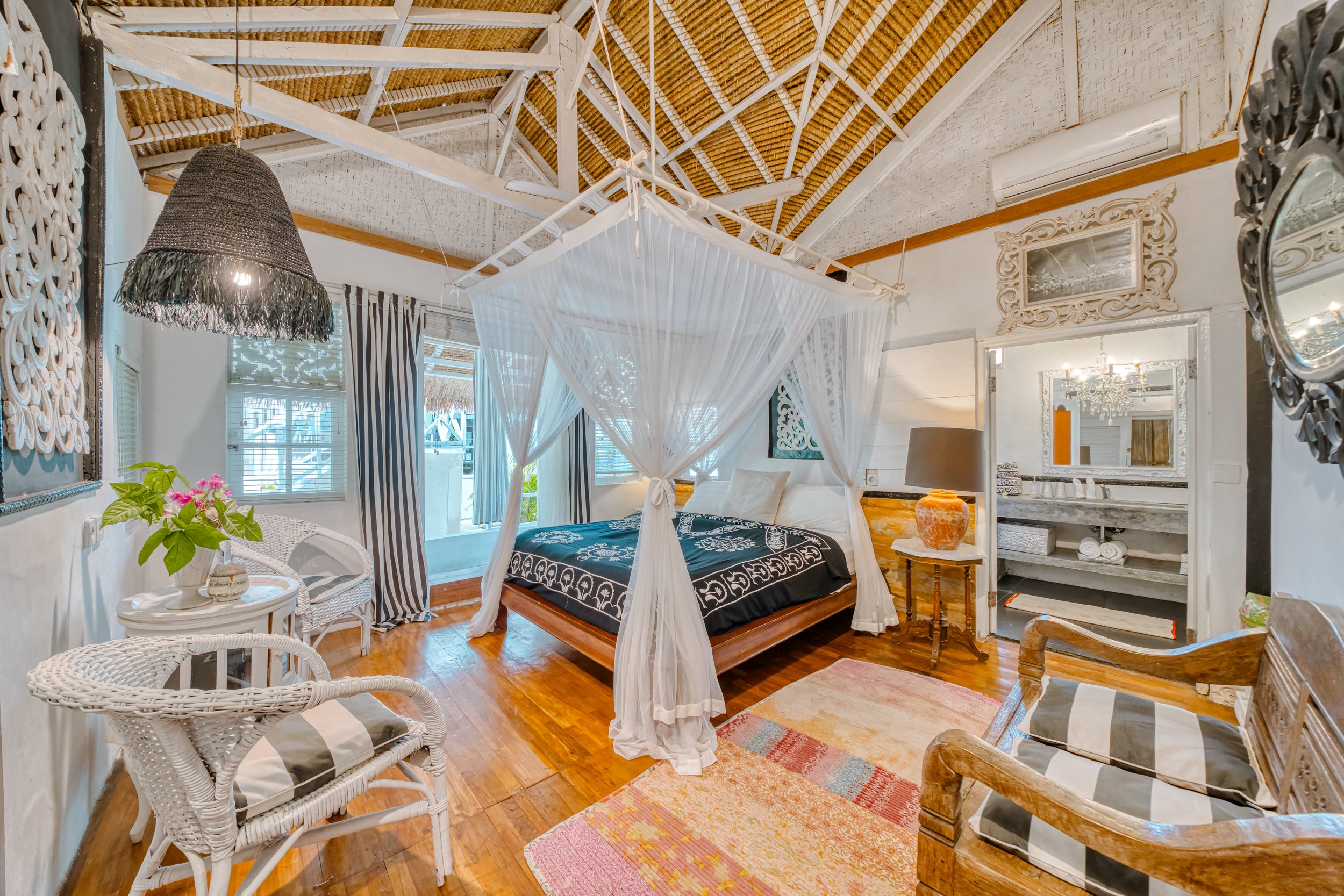 A cozy tropical bedroom with a four-poster bed, white curtains, wood and white walls, wooden ceiling with exposed beams, wooden furniture, striped curtains, and colorful rugs.