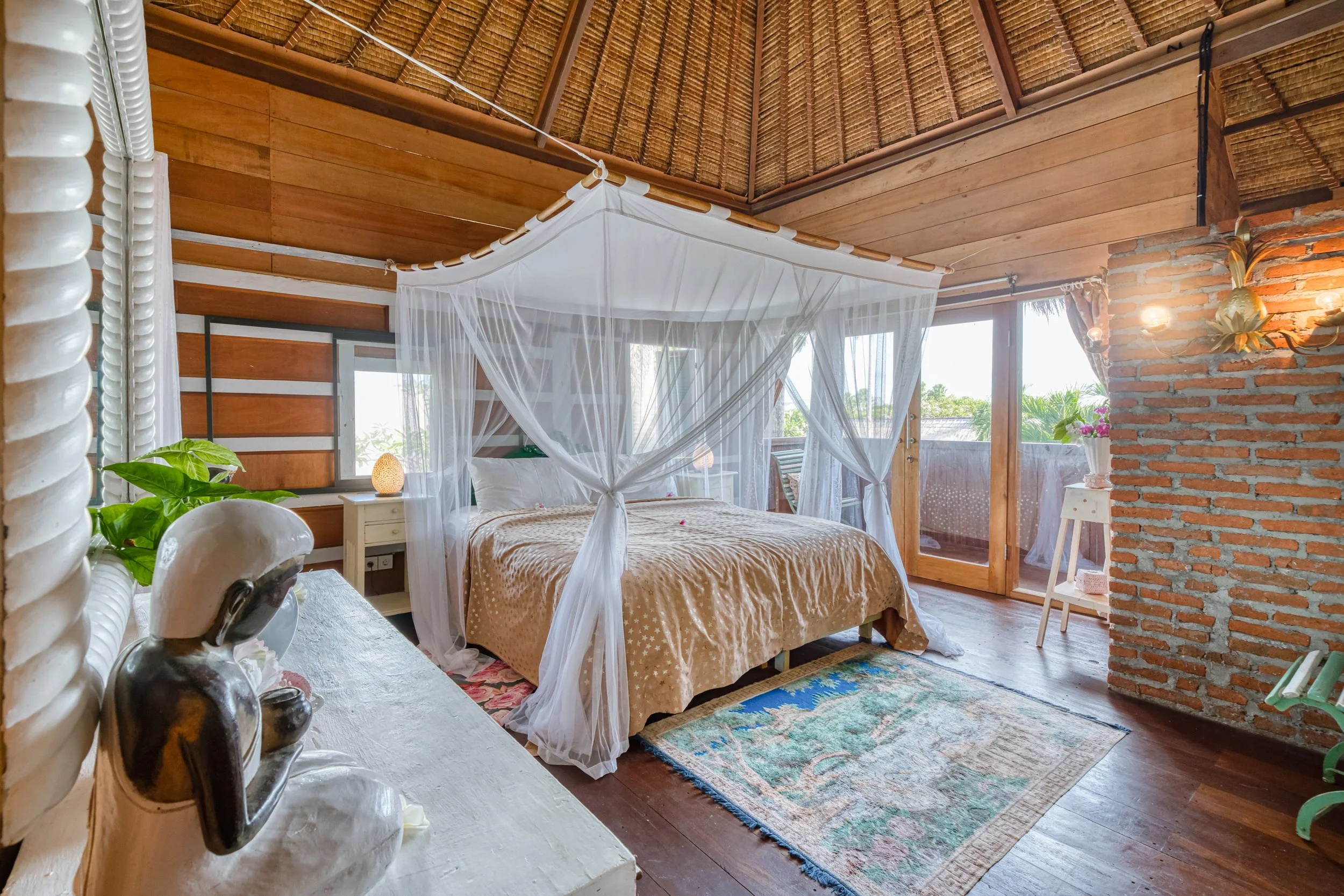 A bedroom with a four-post bed with white sheer curtains, wooden walls, a brick wall, a small white side table with a lamp, a balcony with a sliding door, and tropical plants outside.