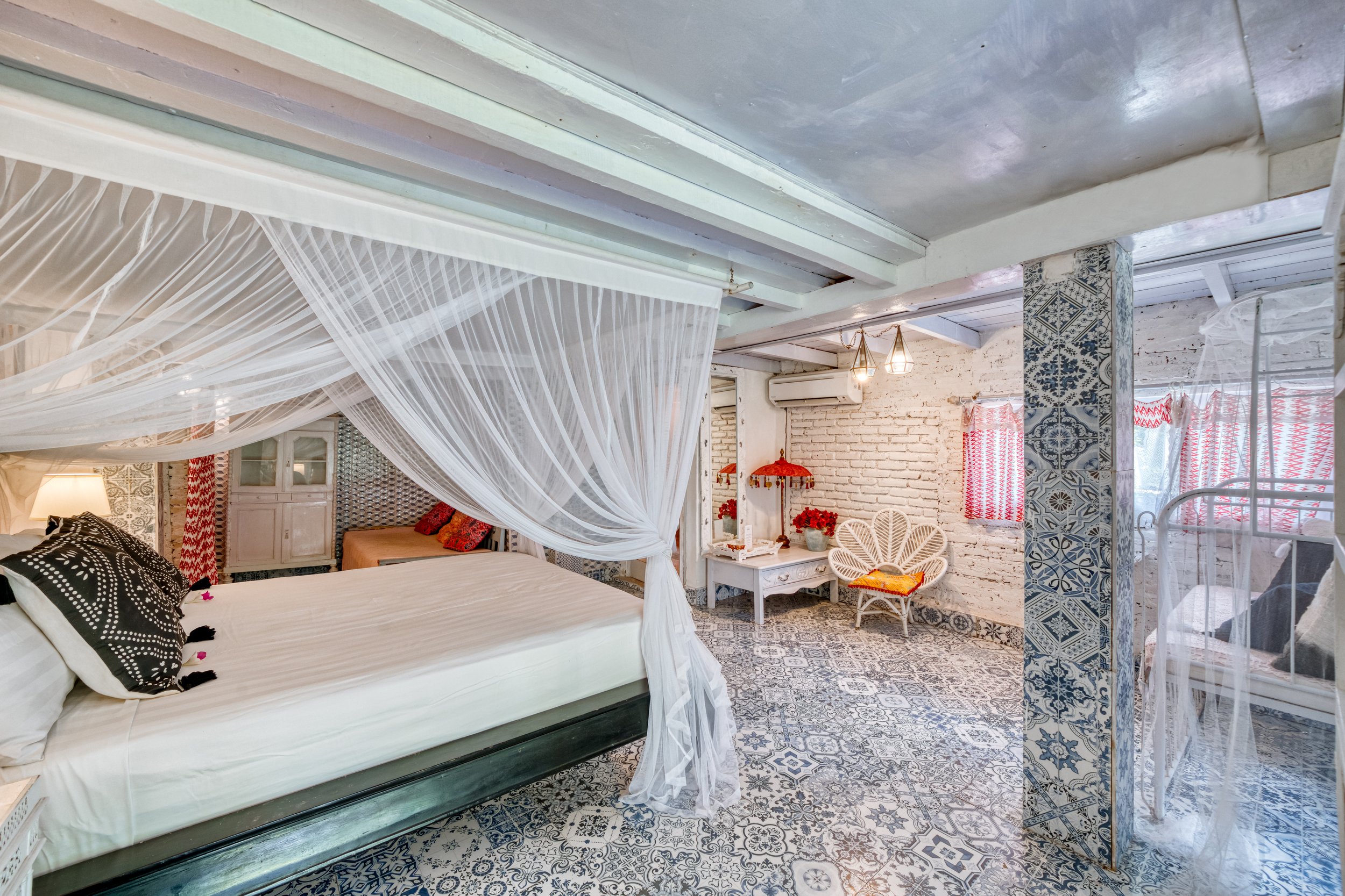 A bedroom with a white canopy bed and black and white patterned pillows, decorated with sheer curtains, a small sitting area with a white wicker chair, and red accents including curtains and flowers on a white table.