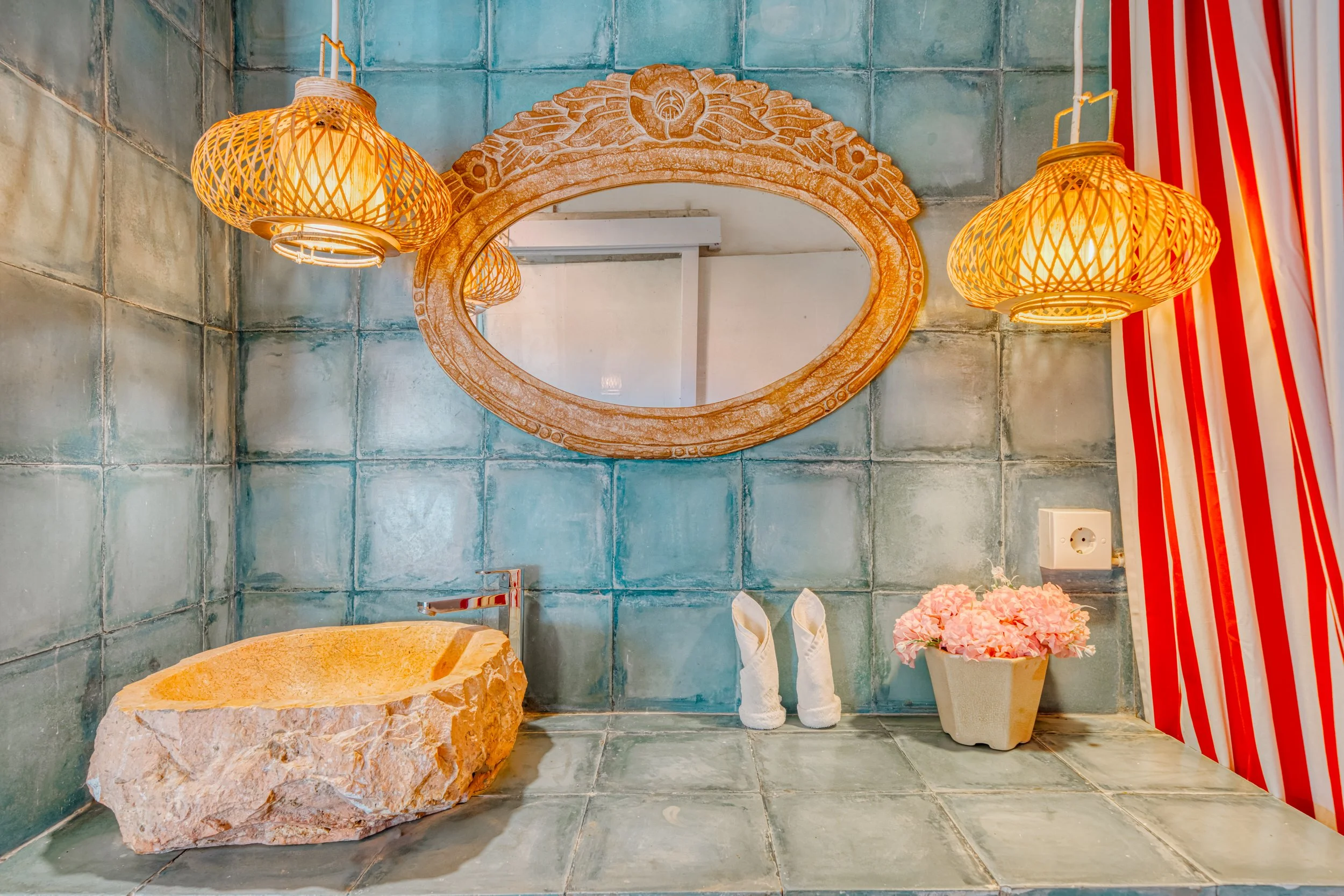 Decorative bathroom with turquoise tiled walls, a large oval mirror, two yellow woven pendant lights, a stone sink, a potted pink flower, and red and white striped curtains.