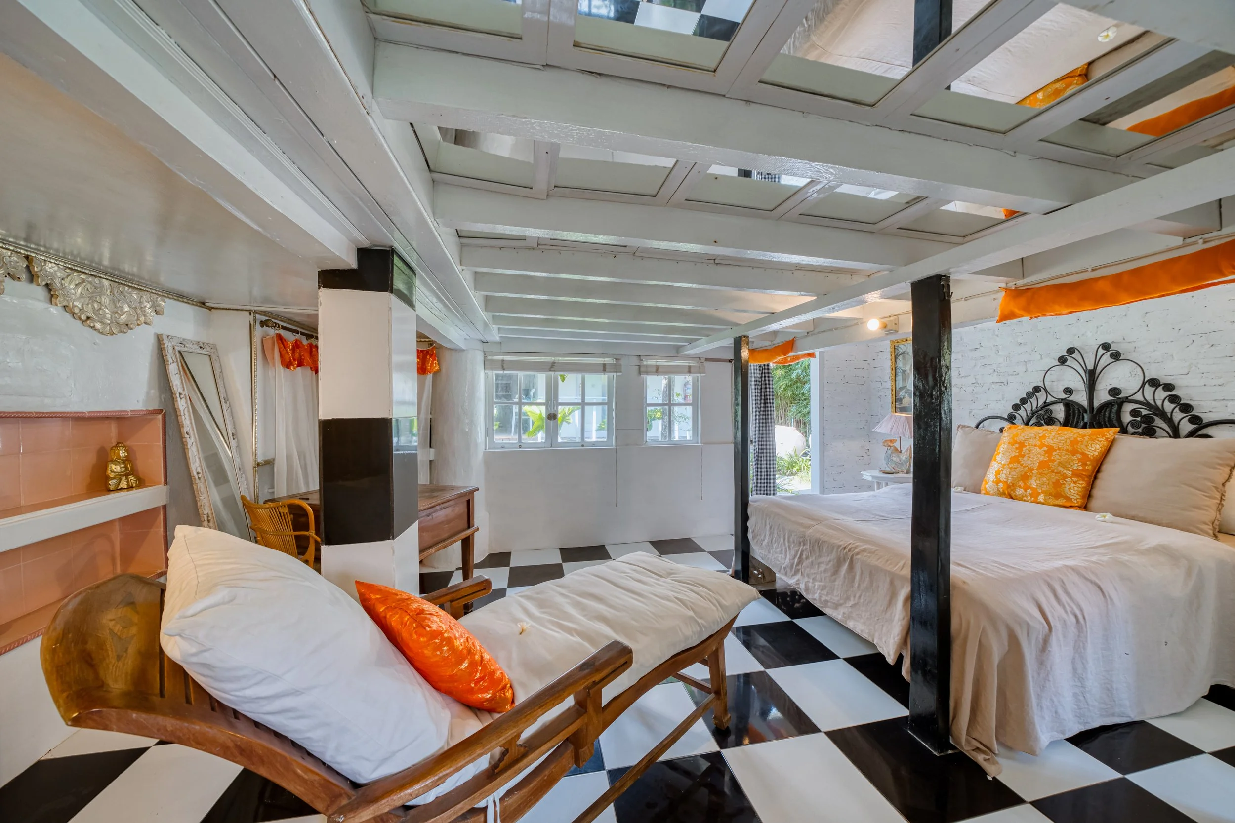 Bedroom with a four-poster bed, orange pillows, and a bench with an orange pillow. The room has black and white checkered flooring, white brick walls, and windows letting in natural light.