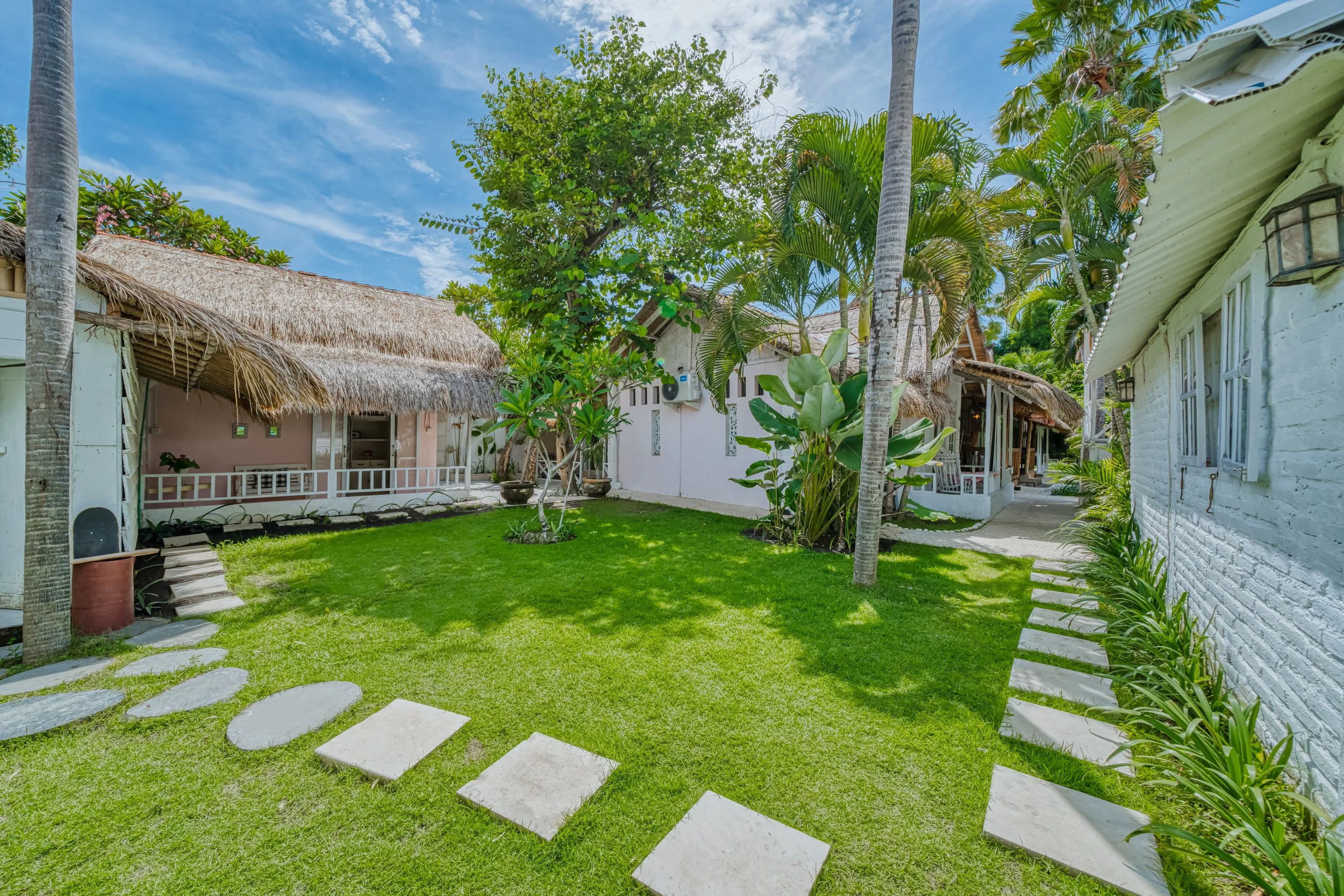 Tropical garden courtyard with green grass, stepping stones, palm trees, and thatched roof cottages under a blue sky.