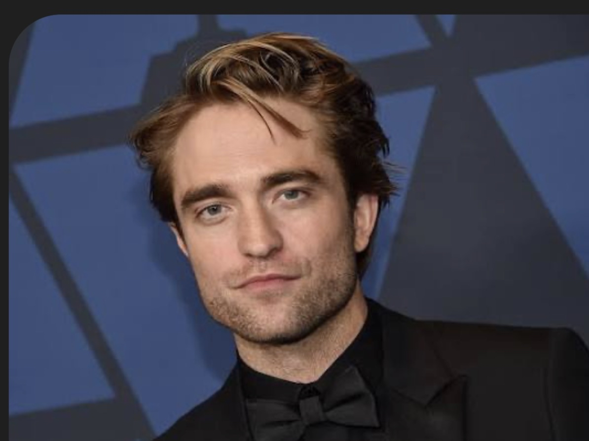 A young man with wavy light brown hair, dressed in a black tuxedo with a bow tie, posing against a dark background.