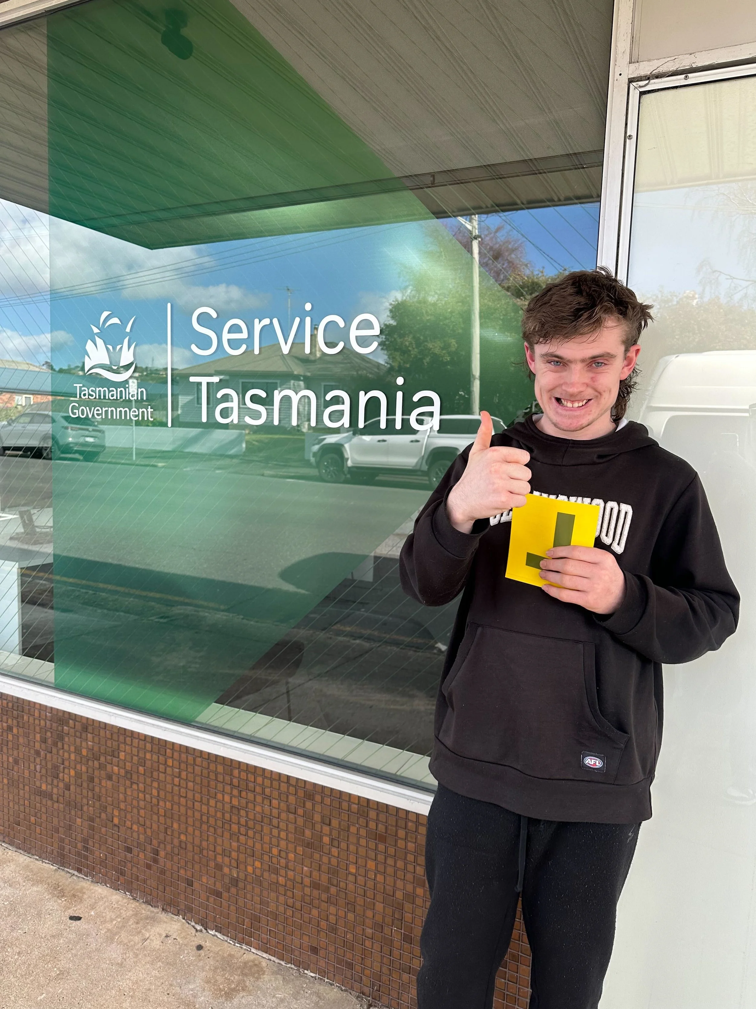 Young man standing outside Service Tasmania window, smiling and giving a thumbs up, holding a yellow document. Reflection of parked cars and trees visible in the window.