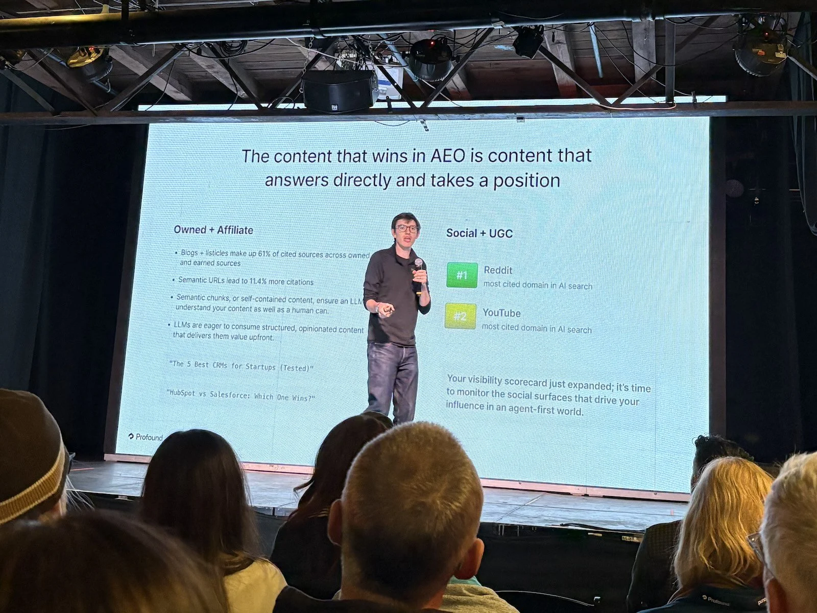A white man with dark hair wearing casual clothes gives a presentation on a stage. The slide behind him reads "The content that wins in AEO is content that answers directly and takes a position."