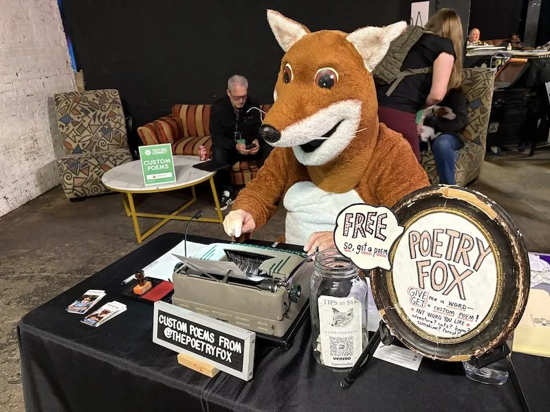 A person in a fox-costume sits at a typewriter and writes a custom poem at a conference.