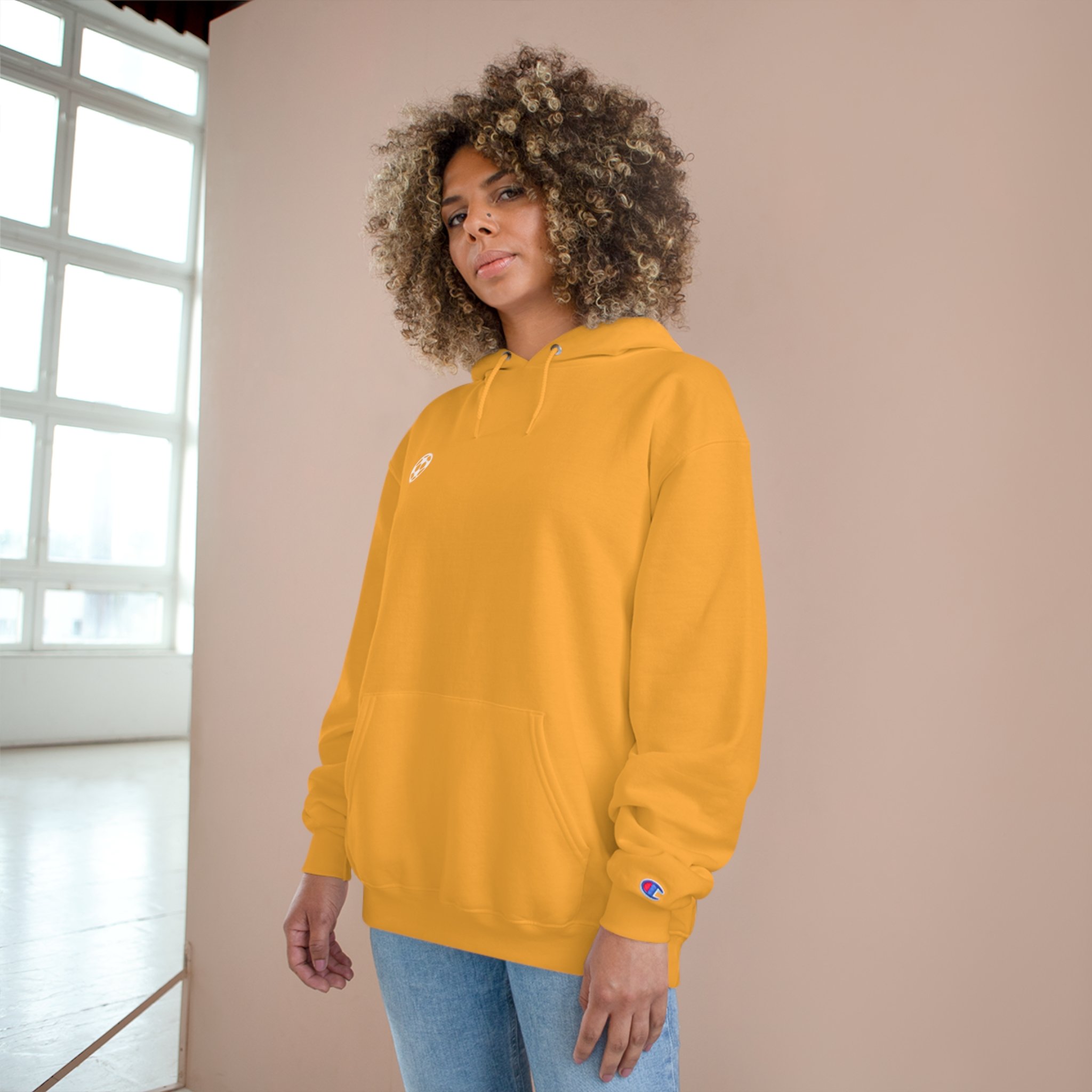 A woman with curly hair wearing a yellow hoodie and light blue jeans, standing indoors near a window.