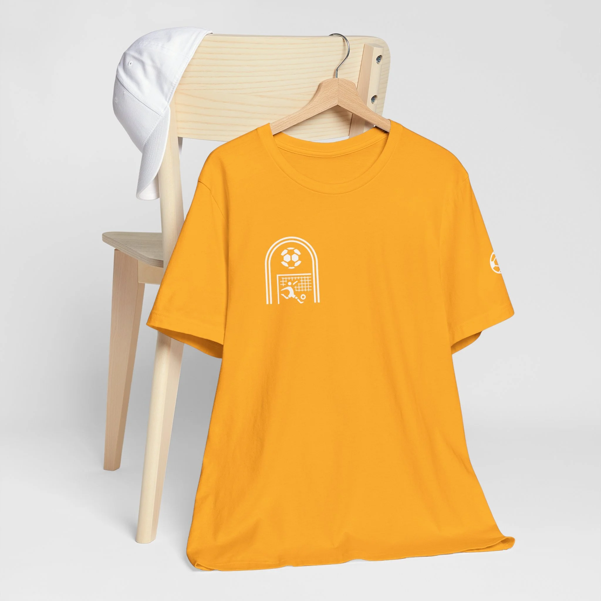 Yellow T-shirt with white soccer-related graphics hanging on a wooden hanger, draped over a wooden chair with a white cap resting on the chair's backrest.