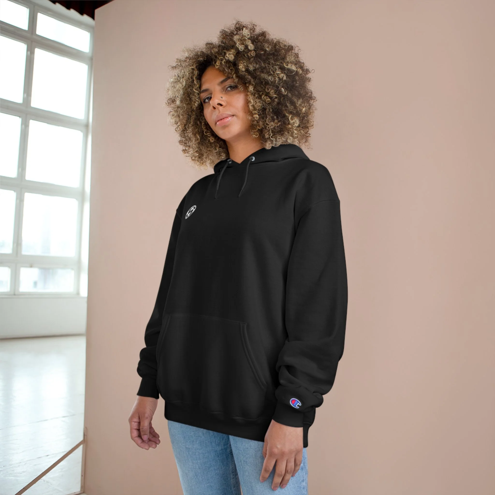 A woman with curly hair wearing a black Champion hoodie and light blue jeans stands indoors against a plain background.